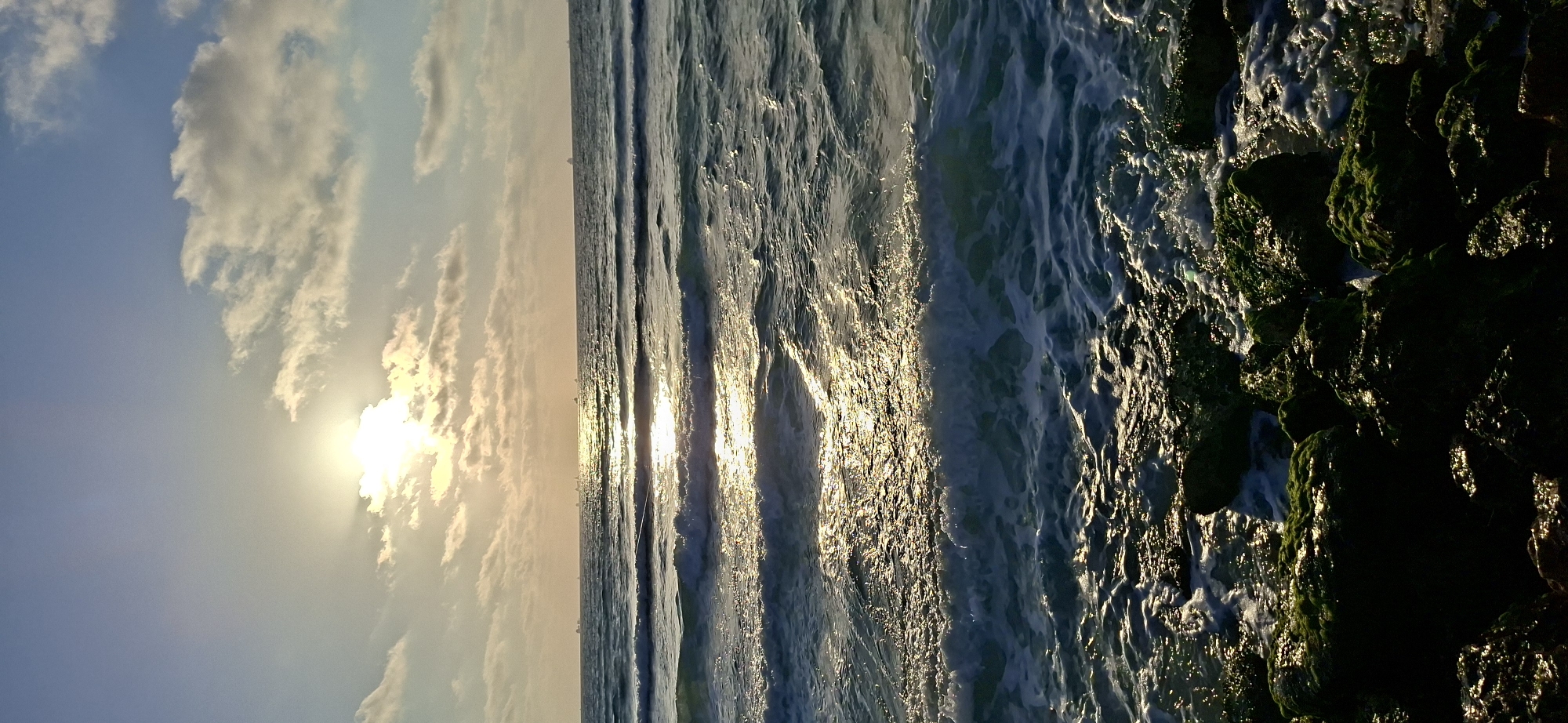 This is a photograph of a ocean scene captured during sunset or sunrise. The sun, a bright orb, is partially obscured by clouds, casting a warm, golden glow across the sky and water. The sky is a gradient of soft blue and pastel hues, with wispy clouds scattered across it. The ocean is a deep blue, with waves gently rolling towards the shore. The sunlight reflects off the water, creating a shimmering path of light that stretches towards the horizon. On the right side of the image, there are dark, moss-covered rocks that are being washed by the waves. The overall mood of the image is serene and peaceful. There is no visible text in the image.