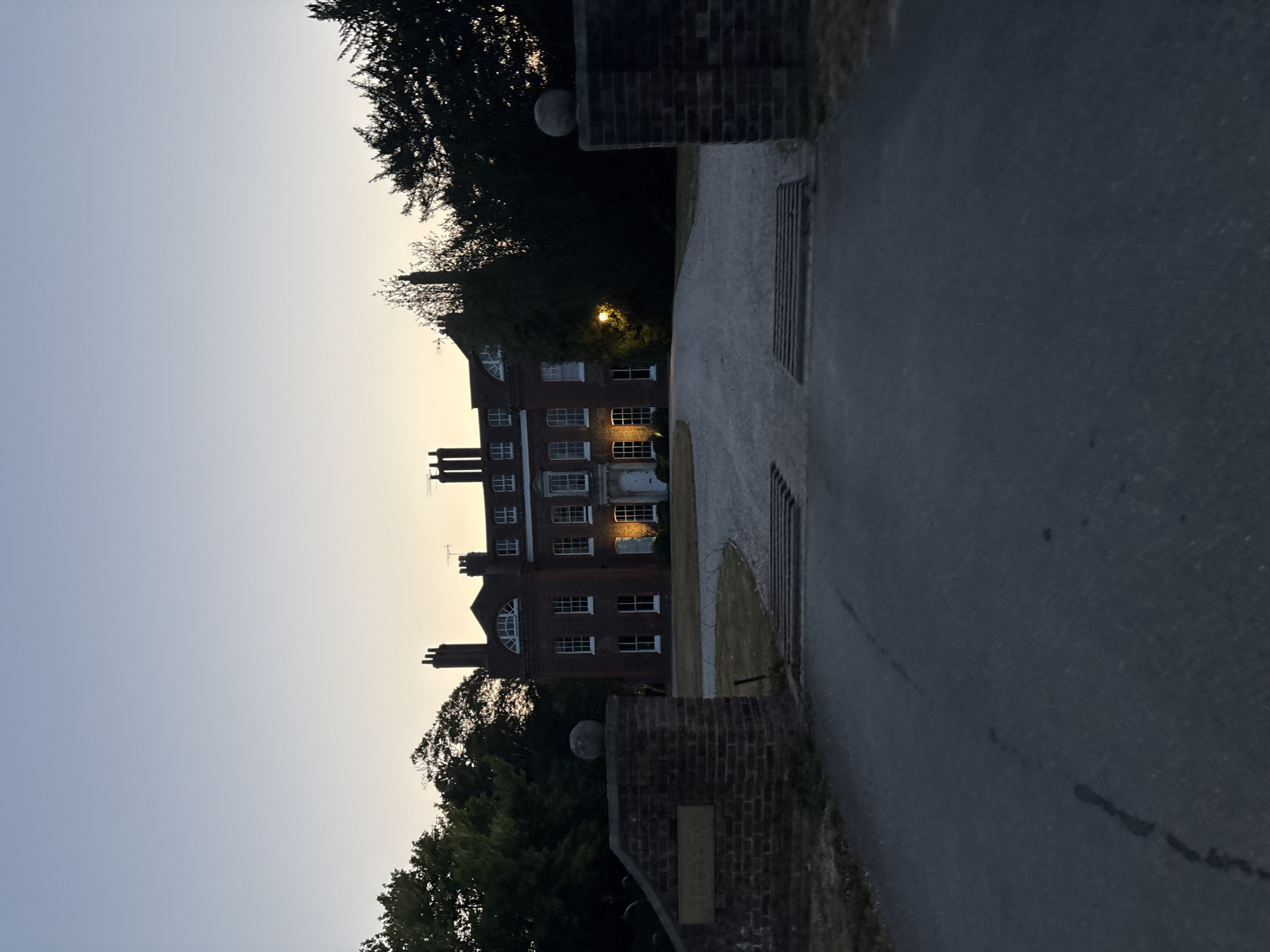 The image shows a large, red-brick house with multiple chimneys, illuminated by interior lights, at dusk. The house appears to be a manor or country house, situated behind a driveway and framed by trees. The foreground features a paved road leading to the driveway, with brick pillars and a signpost on either side. The sky is a gradient of light blue and yellow, indicating the transition from day to night. The scene is devoid of people or any observable activity, suggesting a quiet, possibly private, setting. The presence of mature trees and the architectural style of the house suggest a location in a rural or semi-rural area, potentially on the outskirts of a city like Chelmsford.