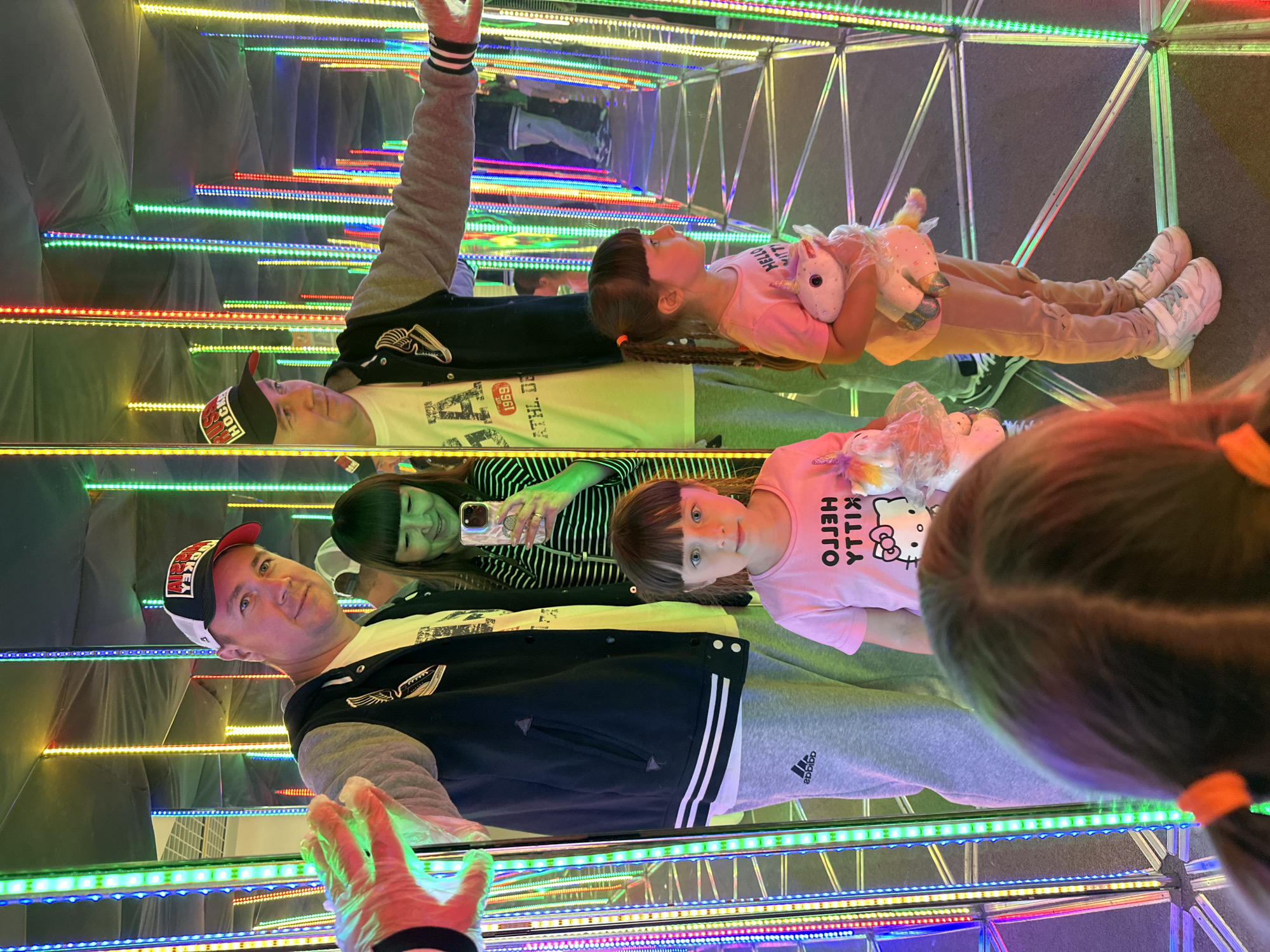 The image shows a family inside a hall of mirrors illuminated by vertical LED strips of various colors. A man in a black and gray jacket and a white t-shirt, wearing a baseball cap, is visible on the left, extending his gloved hand towards the camera. Next to him is a woman in a striped shirt, holding a smartphone. In the foreground, two young girls are present. The girl on the right wears a pink t-shirt with "HELLO KITTY" text and is holding a stuffed unicorn. The girl on the left, partially visible, has her back to the camera and wears a pink t-shirt. The background reflects the family and the colorful lights through the mirrors, creating a repetitive and immersive effect. The overall scene appears to be an amusement or entertainment venue. Based on the provided information, this scene is located in Izhevsk, Russia.