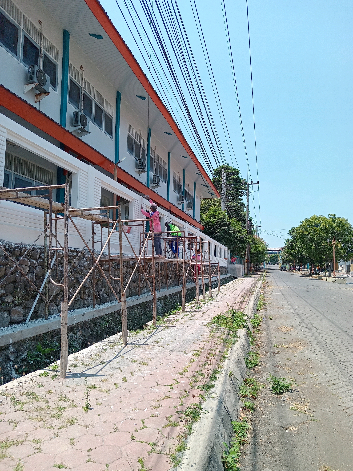 The image depicts a street scene in Randuagung, Indonesia, on a clear day. A two-story building, possibly a school or office, with white walls and orange trim runs along the left side of the frame. Air conditioning units are mounted on the exterior walls. Scaffolding is erected in front of the building, and three individuals are visible on the scaffolding, appearing to be painting or performing maintenance work. One person in a pink shirt is actively painting, while another in a green shirt is standing nearby. A third person, in the background and partially obscured, is also on the scaffolding.
The building overlooks a pathway made of hexagonal pavers, which is lined with a concrete curb and a drainage ditch. The ditch contains some greenery and rocks. Beyond the pathway, a paved road stretches into the distance, with trees lining both sides. Several power lines are strung across the sky above the building and the street. In the background, a vehicle is visible on the road, and a distant structure with an archway can be seen. The overall impression is one of ongoing renovation or construction in a semi-urban environment.