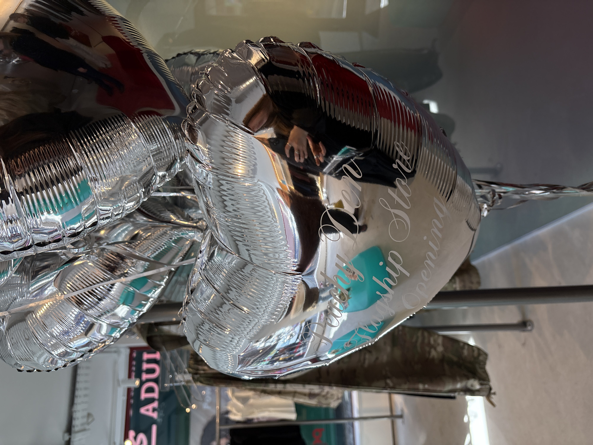 The image shows a close-up of two silver heart-shaped balloons, likely helium-filled, resting on a reflective surface, possibly a glass or glossy table. The balloon closer to the viewer has lettering in a light color, possibly white or silver, which reads "Flagship Opening". The surrounding environment appears to be an indoor retail space, given the presence of clothing and signage. A sign with the word "ADULT" is partially visible behind the balloons. The reflection in the balloon reveals the photographer's hand, holding a phone or camera. The other reflections suggest a busy retail environment.
While not explicitly stated, the lettering on the balloon points to a flagship store opening in London.
