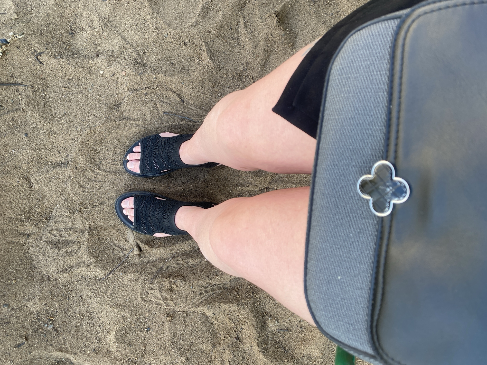 A first-person perspective shot shows a person's legs and feet standing on sand, with a portion of a black and gray handbag in the foreground. The sand appears to have imprints from footwear and possibly some scattered debris like small twigs. The person is wearing black sandals with a woven design and a black skirt or shorts. The lighting suggests it is daytime. The scene is located on the sandy embankment of the Kama River in Chaikovsky, Russia.