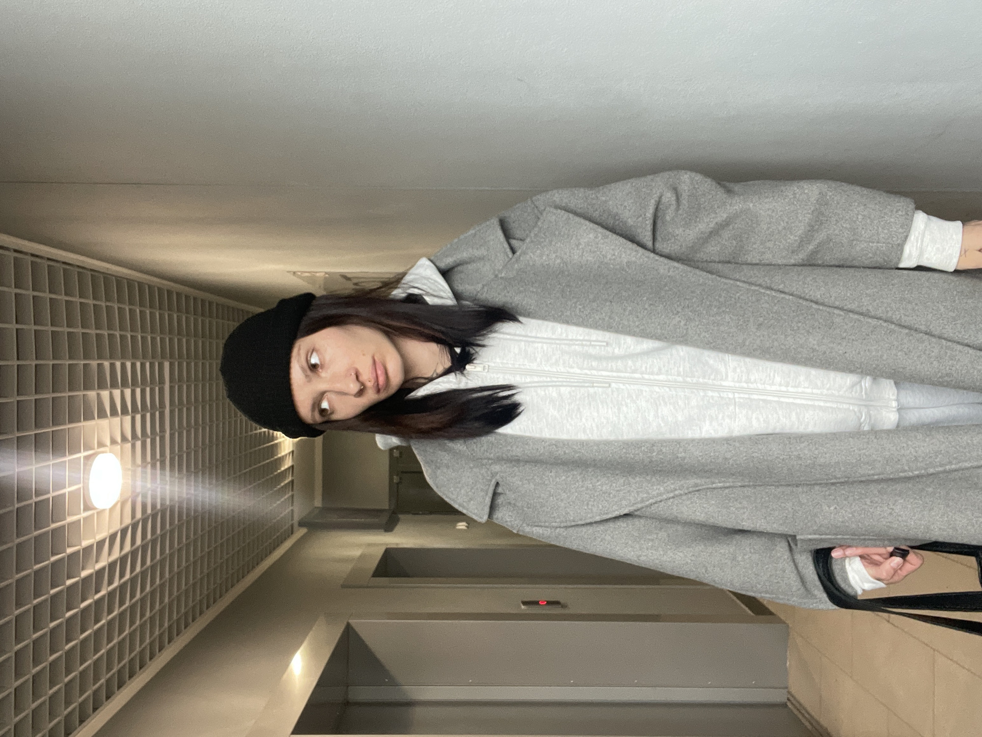 A woman stands in what appears to be an interior hallway or lobby of a building. She is wearing a black beanie, a light grey hooded sweatshirt with a zipper, and a large grey wool coat. She appears to be holding a dark object or bag strap. The wall behind her has a textured, grid-like pattern and is lit by a single, round light fixture. Further down the hallway, there is an elevator and a stairwell. There is no verifiable information about the location of this image in Izhevsk, Russia from the provided image alone.