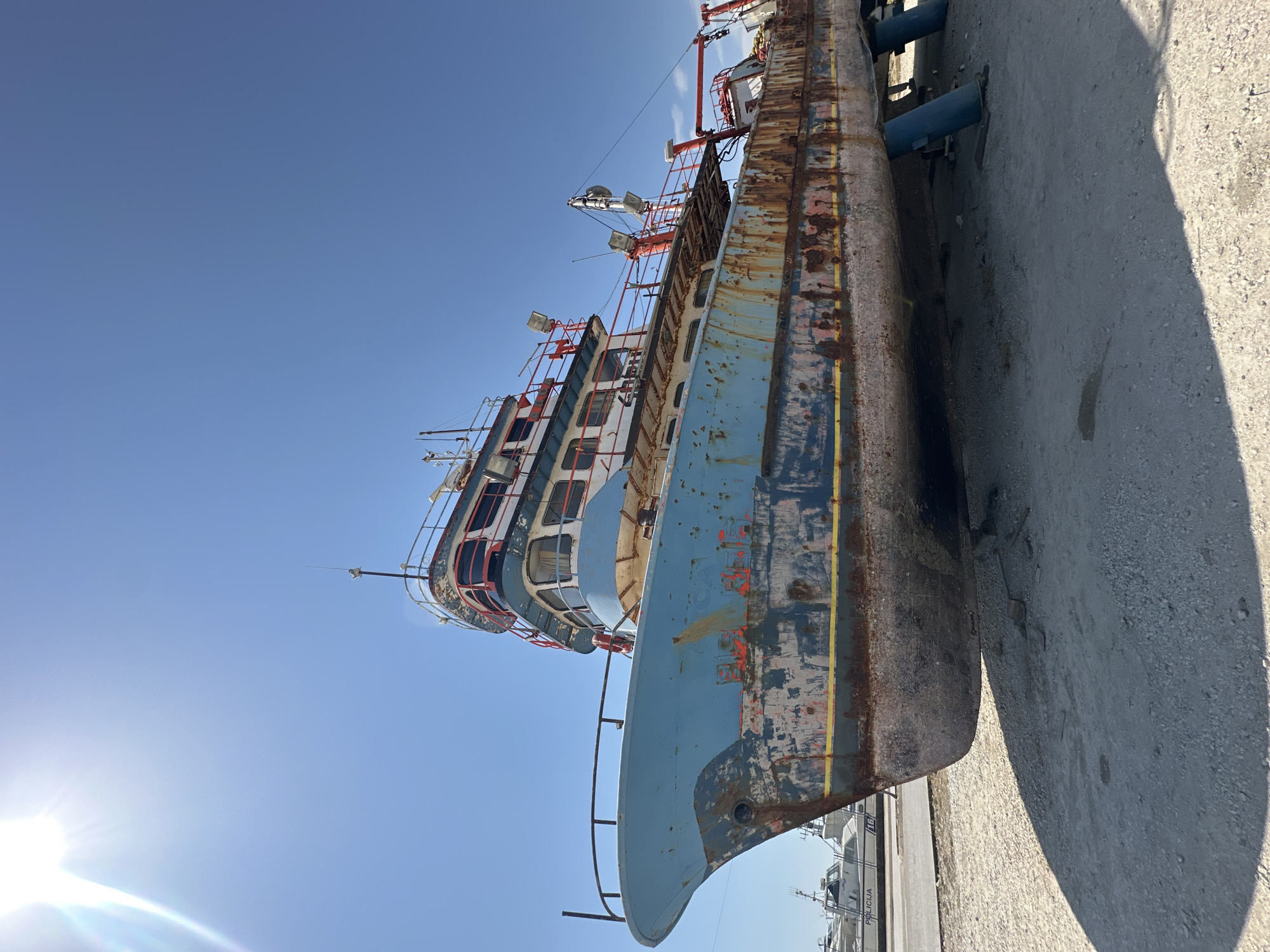 The image shows a large, weathered blue and white ship grounded on a gravel lot under a clear blue sky. The ship, which appears to be a former fishing vessel or similar working boat, exhibits significant rust and peeling paint along its hull, particularly around the waterline. It is propped up on blue stands, indicating it is out of the water. The ship has multiple decks with windows and railings. In the background, other smaller white boats are visible, one with the word "POLICIJA" (police in various Slavic languages) visible on its side, suggesting the location might be a port or shipyard. The sun is bright, casting a strong shadow from the ship onto the gravel. The scene conveys a sense of disuse and aging, possibly in a dry dock or storage area of the port of Bar, Montenegro.