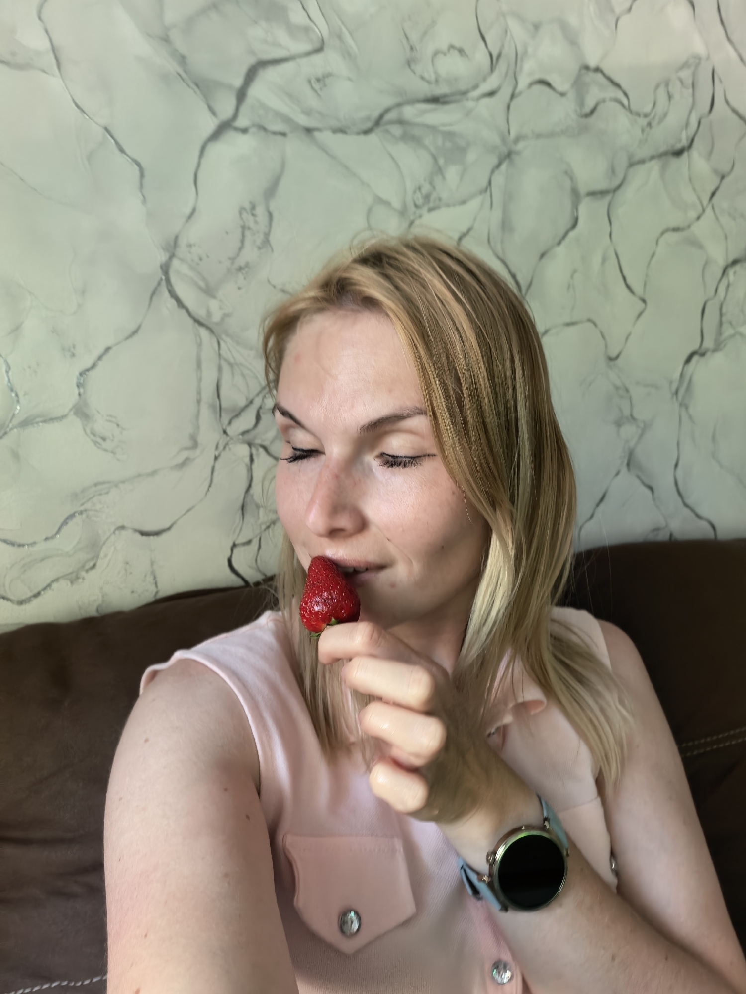 A woman with fair skin and blonde hair, wearing a pink sleeveless top and a smartwatch, is holding a strawberry to her lips with her right hand. Her eyes are closed, and she appears to be smelling the fruit. She is sitting on a brown couch in front of a wall with a marbled pattern. The city is Unknown, Unknown.