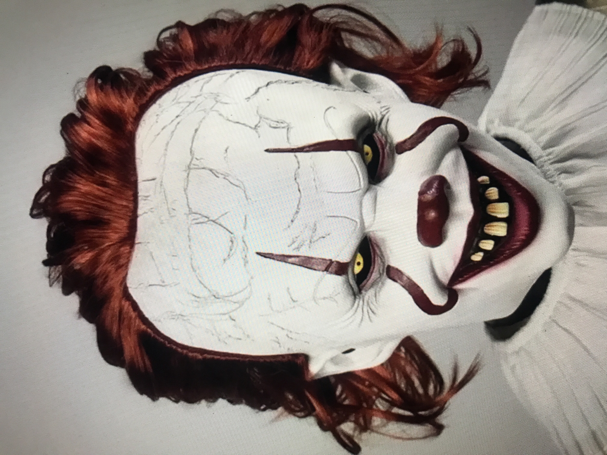 The image shows a close-up of a Pennywise the Clown mask, tilted slightly to the right. The mask has a white face with crack-like markings, yellow eyes, and a wide grin revealing yellowed teeth. It features reddish-brown hair and a white ruffled collar. The background is a plain, light-colored surface. The location in Donji Pobori, Montenegro is unknown.