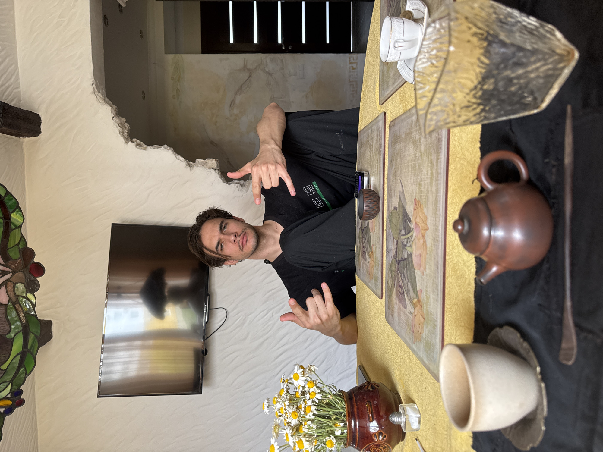 A sideways, indoor shot shows a man standing next to a table laid with items and behind him a doorway leading into another room.
The man is fair-skinned with dark hair. He is wearing a black t-shirt and is making a hand gesture. He is looking directly at the camera. Behind him on the wall, there is a large flat screen television. To the left, is a stained glass fixture and a brown wooden beam.
On the table are various items including a small brown teapot, a white cup, a glass decoration, some coasters, and a small wooden bowl. In the foreground is a vase of white and yellow daisies. The table is covered in a gold textured fabric.
Behind the man is a doorway, with rough edges, leading into another room. The room is lit with white light. In the distance, there are dark wooden doors with vertical LED lighting features.
The city of Sevastopol, Ukraine, is not discernable in the image.