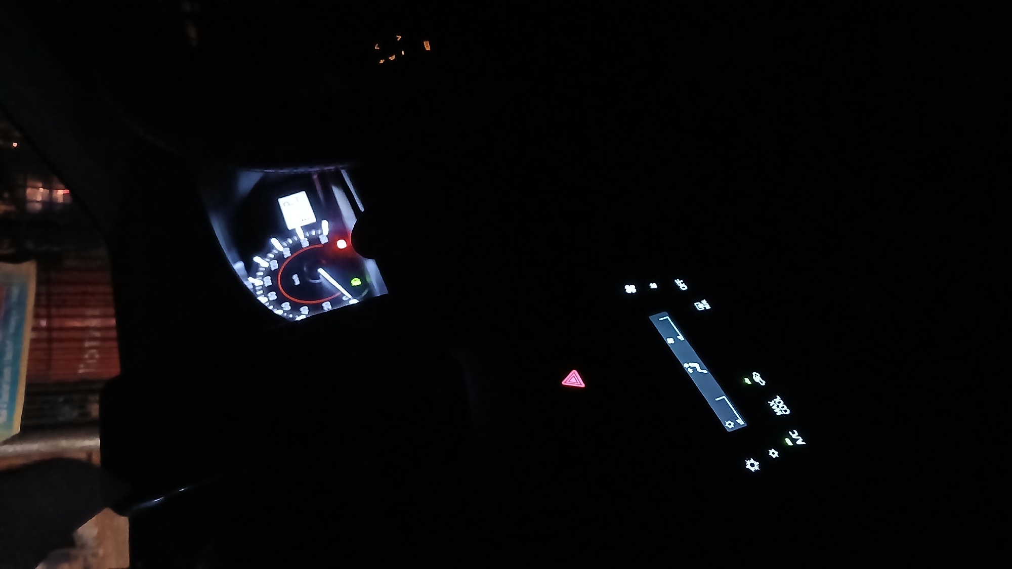 The image shows the interior of a car at night. The dashboard is visible with illuminated gauges, including a speedometer and various indicator lights. The speedometer needle is near zero, and a red warning light is on. The hazard lights button is illuminated. The car's climate control panel is also lit up, showing settings for temperature and fan speed. Outside the car window, blurred lights are visible, suggesting an urban environment. The location is consistent with the city of Lhokseumawe, Indonesia. No people are visible in the image.