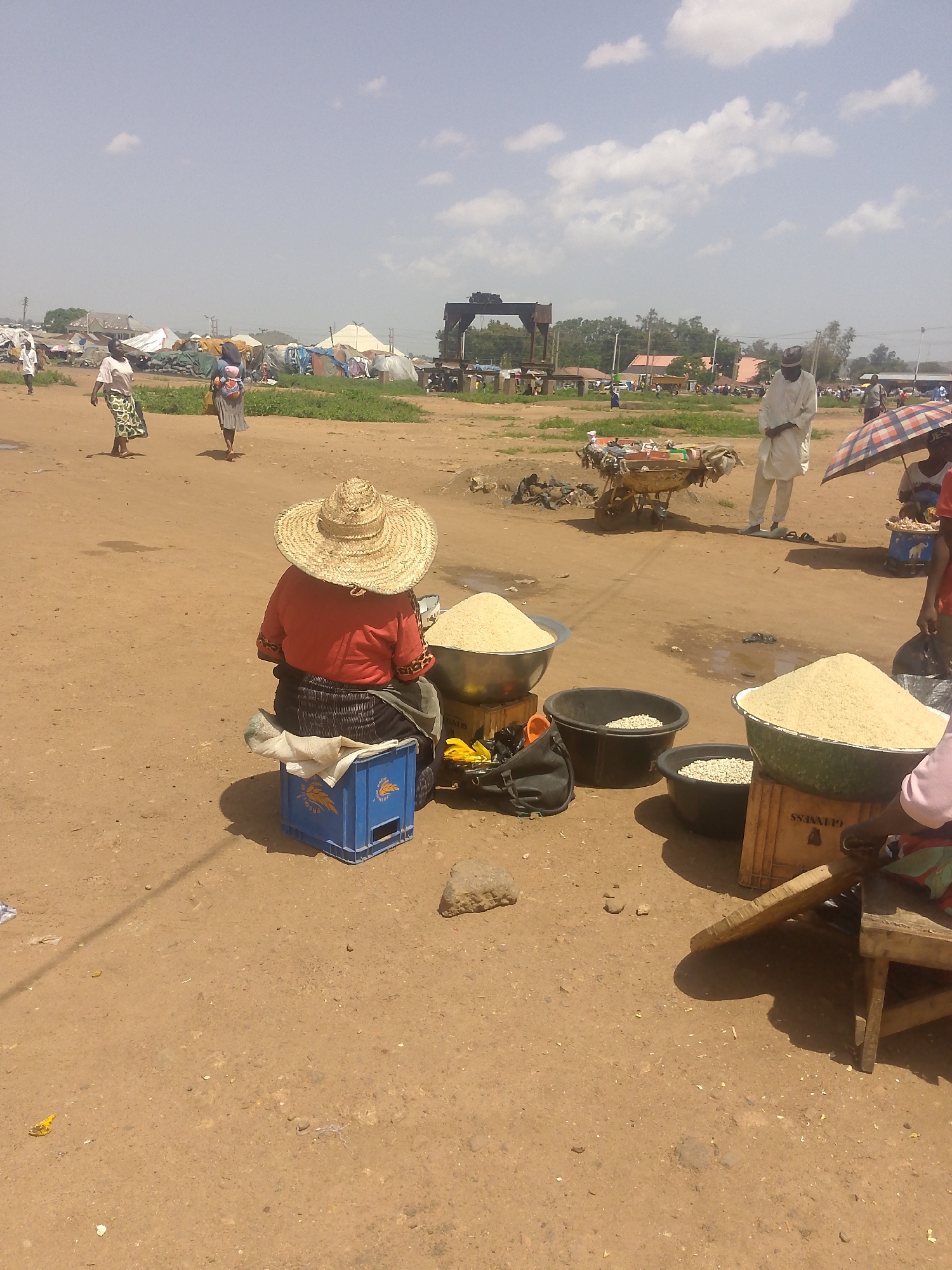 The image captures a market scene on a dirt ground in Unknown, Unknown. In the foreground, a person wearing a straw hat and red shirt sits on a blue crate. Beside them are bowls filled with grains or beans. Other people are present, including a man in white clothing and a woman holding an umbrella. In the background, tents and other market structures are visible.
