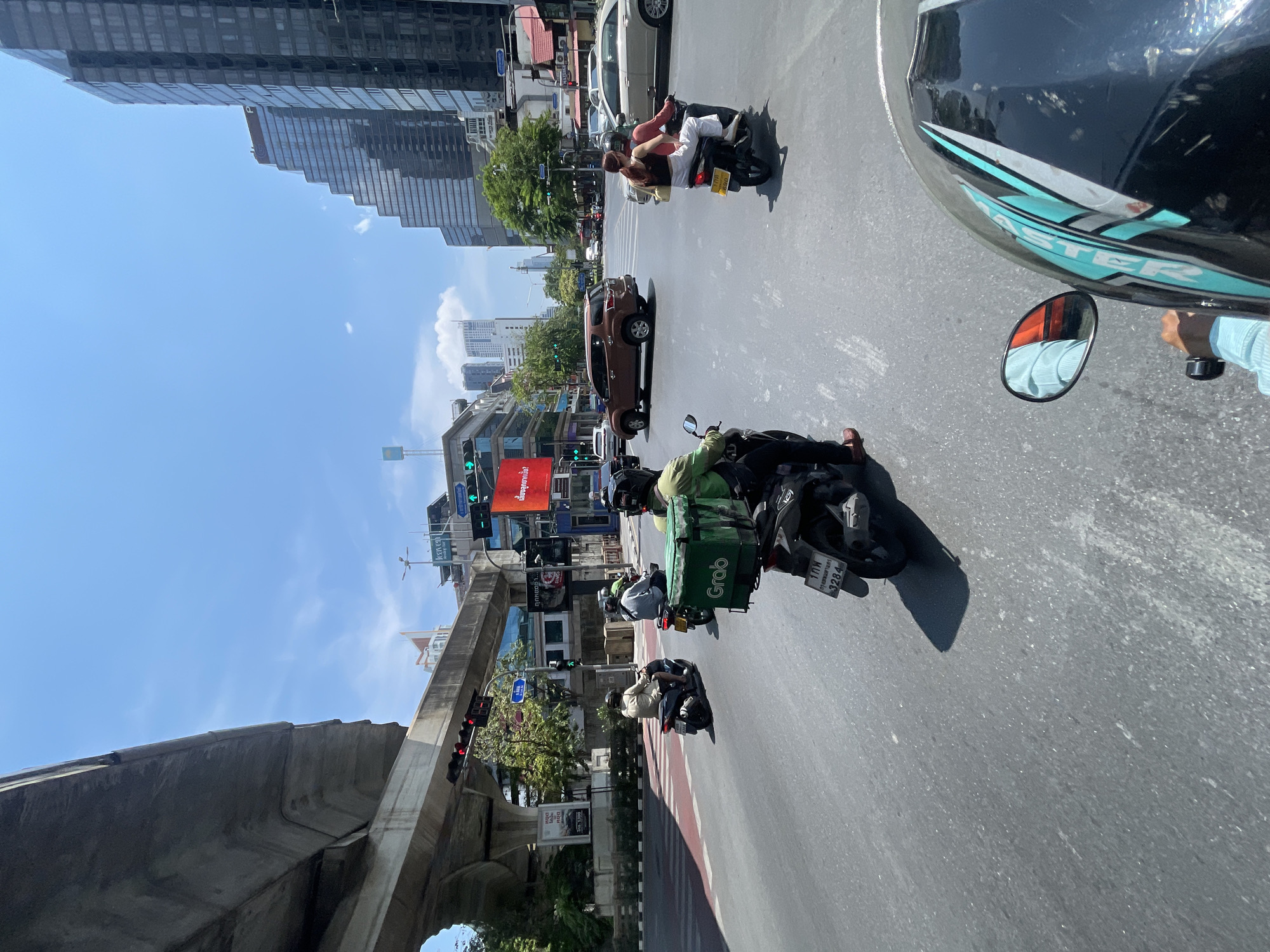 The image captures a street scene in Bangkok, Thailand. The perspective appears to be from a moving vehicle, likely a motorcycle. The focus is on the urban environment with several motorcycles navigating the street. A delivery motorcycle with a green "Grab" delivery bag is prominently displayed. The Grab motorcycle registration reads "1 กก กรุงเทพมหานคร 3284". A couple on a motorcycle is close by, as is a brown SUV. A large red advertising screen hangs above the road. An elevated train track runs above the street, with visible signage and traffic signals. Skyscrapers and other buildings form the backdrop, indicating a bustling city center. The sky is clear and blue, suggesting a sunny day.