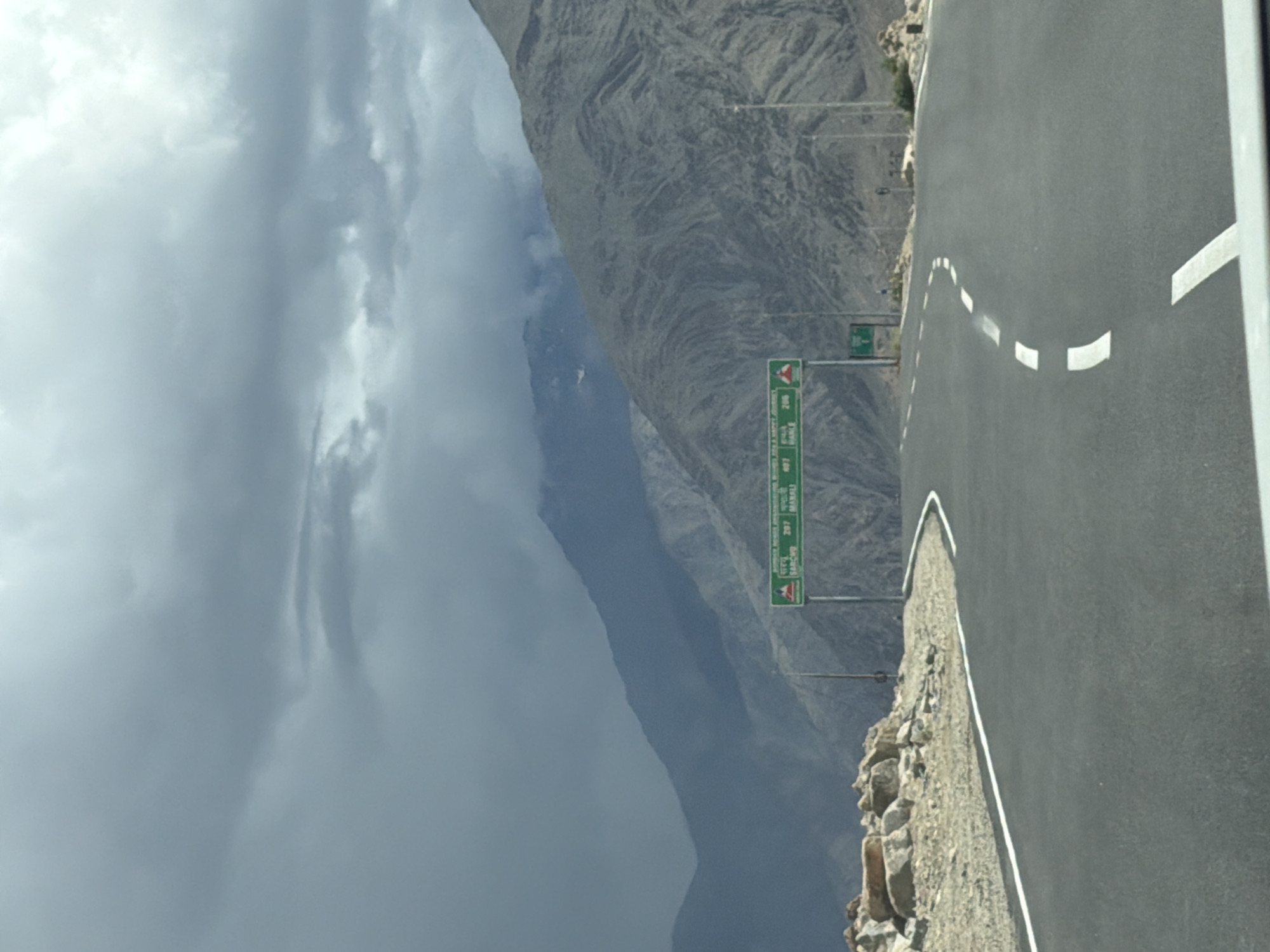 This image captures a scenic view of a winding asphalt road in Upshi, India, a region known for its mountainous terrain. The road, marked with white lines, curves gently through a landscape dominated by rugged, barren mountains. Dark, ominous clouds gather overhead, suggesting the possibility of rain or stormy weather, creating a dramatic atmosphere. On the right side of the road, a green directional signpost displays distances to various locations, including Sarchu (297 km) and Manali (407 km). Rocky outcrops and sparse vegetation dot the foreground, further emphasizing the wild, untamed nature of the environment. No people or vehicles are visible in the frame, giving a sense of solitude and vastness to the scene. The overall impression is one of a remote and awe-inspiring natural setting, with the road serving as a pathway through the imposing mountains.