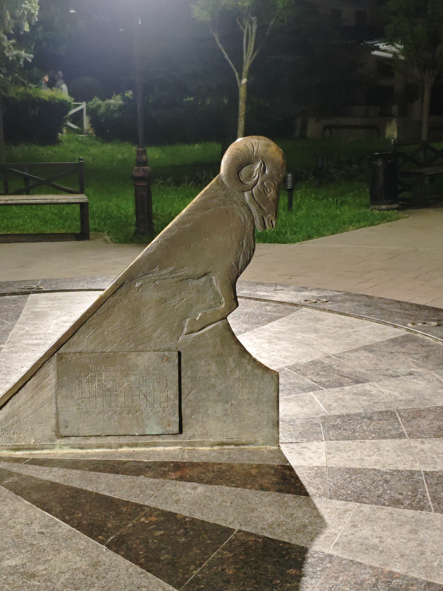 The image captures a sundial sculpture featuring a ram's head in a park setting in Pyatigorsk, Russia. The sculpture is bronze-colored and includes a plaque with "The Equation of Time" inscribed above a graph. The sundial is positioned on a tiled surface with a radiating pattern. In the background, there is a park area with benches, a lamppost, trees, and some people visible in the distance. The scene is lit by artificial lighting, suggesting it is nighttime.