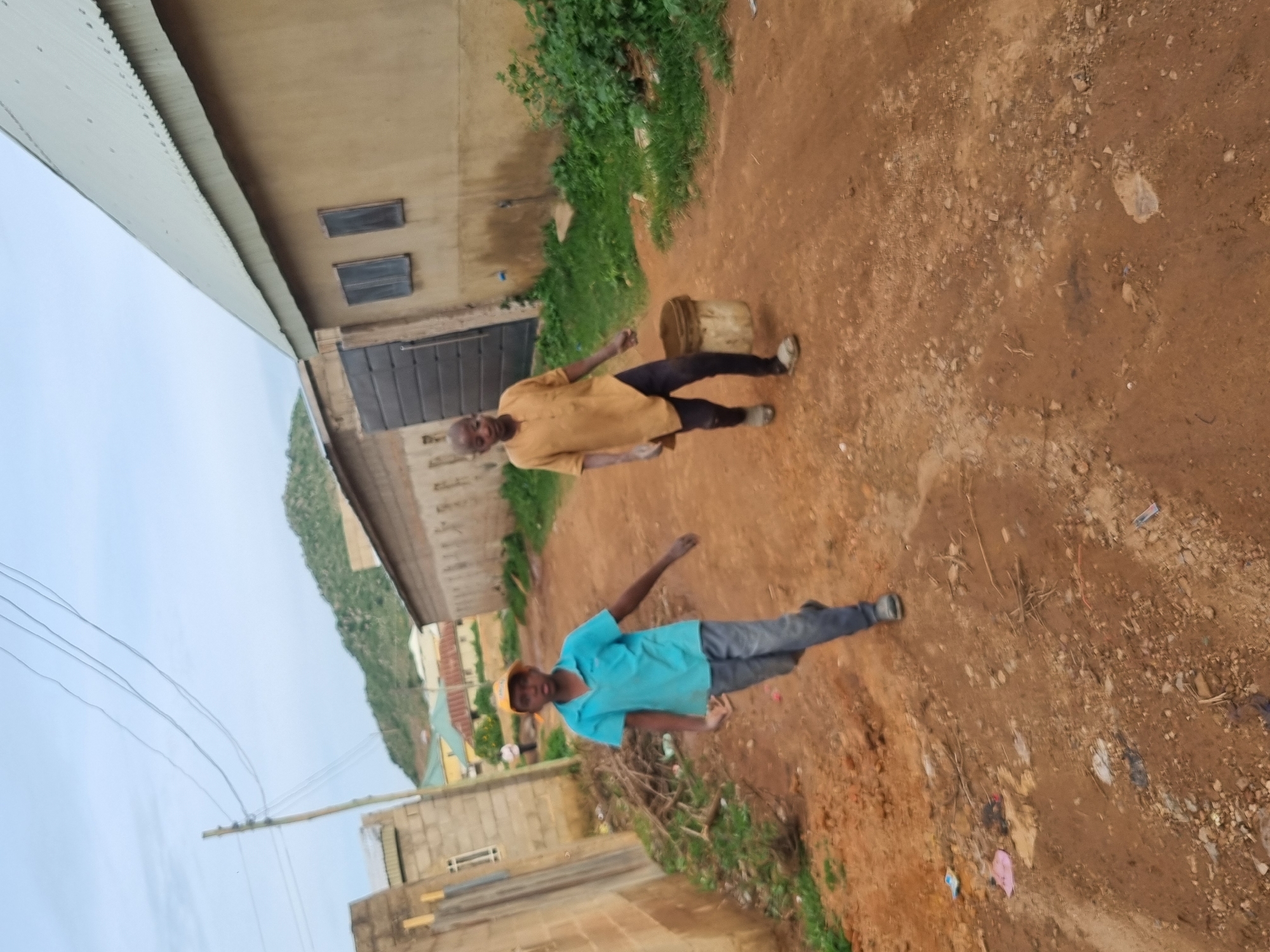 The image shows two men standing outdoors in what appears to be a rural or semi-urban area in Jos, Nigeria. The ground is a reddish-brown dirt, common in such locations. To the left, a light-colored building with a corrugated metal roof is visible, along with some windows and a large metal door. In the background, there is lush green vegetation and some other buildings.
The man in the foreground is wearing a bright blue t-shirt and dark pants. He is looking towards the camera with a neutral expression. The second man, positioned further back and to the right, is wearing a yellow shirt and dark pants. He is also looking towards the camera, and his right arm is slightly raised. A light-colored bucket is visible on the ground near him.
The scene seems to be an everyday moment, possibly during the daytime given the natural light. The presence of the buildings, the dirt ground, and the vegetation suggests a location that is not a highly developed urban center. There is no visible text in the image. The overall impression is one of a simple, outdoor setting with two individuals engaged in an unspecified activity.