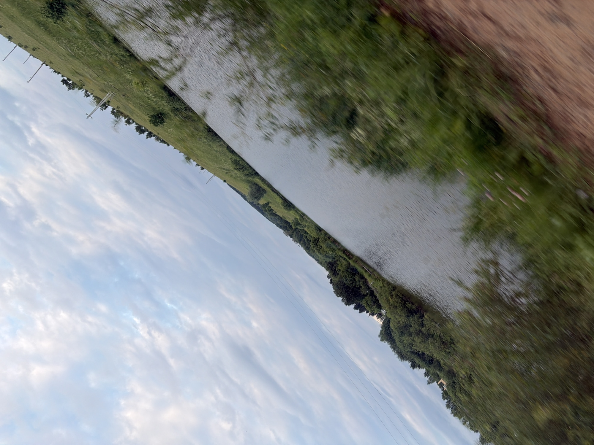 This media file depicts a water body with a grassy embankment on the left and a wooded area on the right, seen from a slightly elevated perspective, possibly from a moving vehicle, indicated by the motion blur. The scene is dominated by natural elements: a wide expanse of water, a green, sloping field dotted with sparse trees and utility poles, and a dense line of trees along the far bank of the water. The sky is partly cloudy with a mix of white and gray clouds against a pale blue background. The orientation of the image is skewed, suggesting it was captured at an angle.
The subjects are the natural landscape itself. The objects visible are utility poles and wires running along the grassy embankment. The scene is a rural or semi-rural setting, with a body of water that could be a river, lake, or pond. The overall impression is one of a tranquil, natural environment.
There is no discernible activity or event taking place, other than what might be inferred from the motion blur, suggesting travel.
Notable details include the time of day, which appears to be late afternoon or early evening, given the soft lighting and the cloud formations. The weather seems temperate. The presence of utility poles provides a subtle cue of human infrastructure within the natural landscape. There is no visible text in the image. The feeling conveyed is one of peaceful observation of nature.