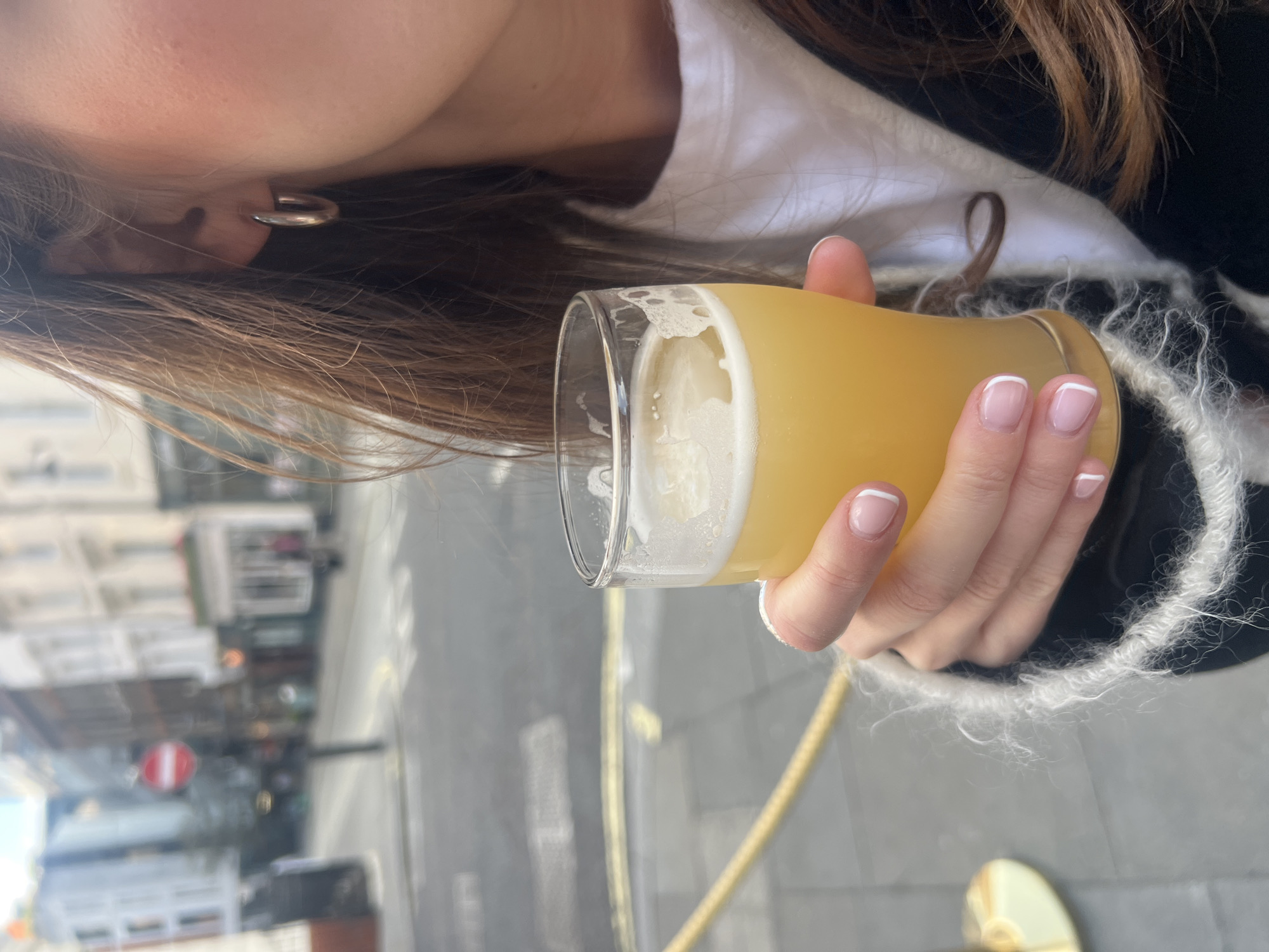 A woman is holding a glass of beer in her right hand outdoors in London, United Kingdom. She has fair skin and brown hair, and is wearing silver hoop earrings. She wears a white shirt under a black and white jacket. Her nails are manicured with a French tip design. The beer is a light color with a foamy head. The background shows a blurred city street with buildings and a "no entry" sign. A golden bar is on the right-hand side.