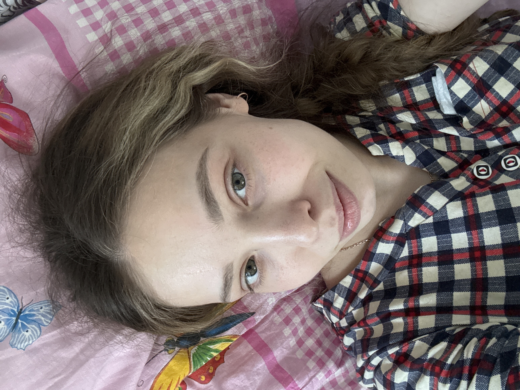 A young woman is lying on a pink and white checkered bedspread. The bedspread is adorned with images of butterflies. She is wearing a red, white, and blue plaid shirt. She has light brown hair and green eyes. She is wearing a gold necklace and small gold hoop earrings. Her location cannot be determined based solely on the provided image. There is no information indicating the presence of Кемуль, Russia, or any other city.