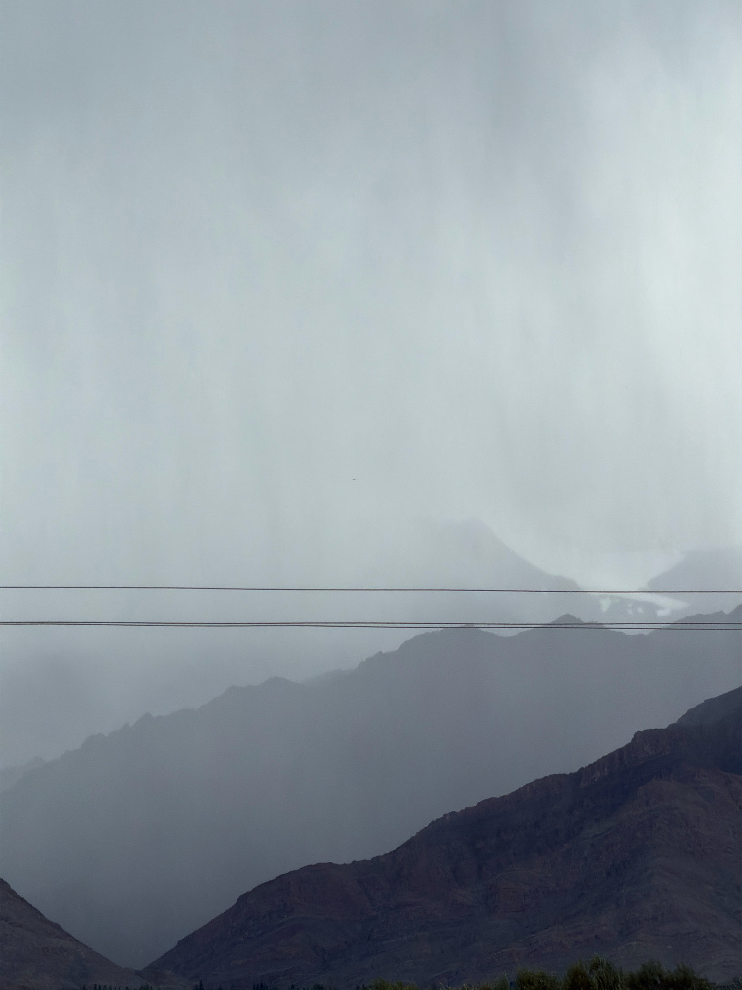 The image captures a desolate, mountainous landscape with a thick, overcast sky. Three horizontal power lines stretch across the middle of the frame, bisecting the view of layered, hazy mountain ranges. The mountains themselves are rendered in muted tones of grey and dark brown, with the foreground peaks appearing more defined and reddish-brown in color. The atmosphere is heavy and somber, suggesting a cloudy or possibly rainy day. There are no people, animals, or man-made structures visible other than the power lines, which themselves are thin and unobtrusive against the vastness of the terrain. The distant peaks fade into the cloudy sky, creating a sense of depth and scale. The overall impression is one of natural grandeur, but with an isolating and somewhat melancholic mood. The location is identified as Yuluk, India.