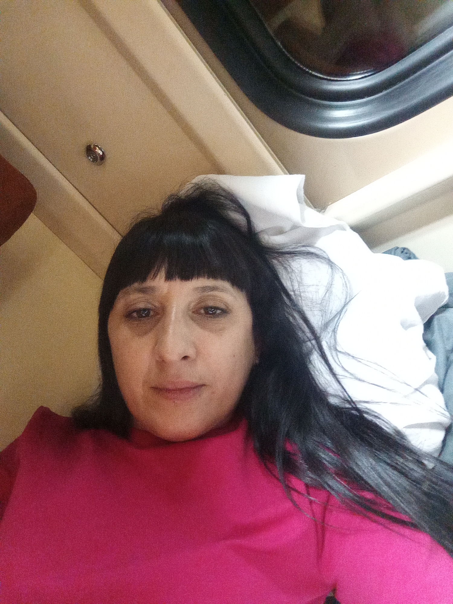 A woman with dark hair and bangs is lying down on a bed in what appears to be a train compartment. She is wearing a bright pink shirt. The compartment has beige walls and a dark-rimmed window. A white pillow and a blue blanket are visible on the bed. There is a small silver and red decorative object on the wall above her.
