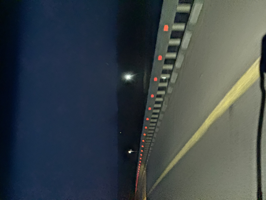 The image displays a nighttime view of a road in Khanskaya, Russia. A two-lane asphalt road, marked by a continuous yellow line on the left, stretches into the dark distance. On the right side of the road, a concrete guardrail features regularly spaced red reflective markers along its upper edge, and alternating black and white vertical sections below. Two distant, bright white artificial light sources are visible against the dark background: one on the left side of the road and another further right beyond the guardrail. The horizon and background beyond the road are obscured by darkness, appearing as indistinct dark terrain. The sky is a deep blue-black. A dark, curved reflection is present at the bottom of the frame, indicating the perspective from inside a vehicle. No individuals or active vehicles are visible.
