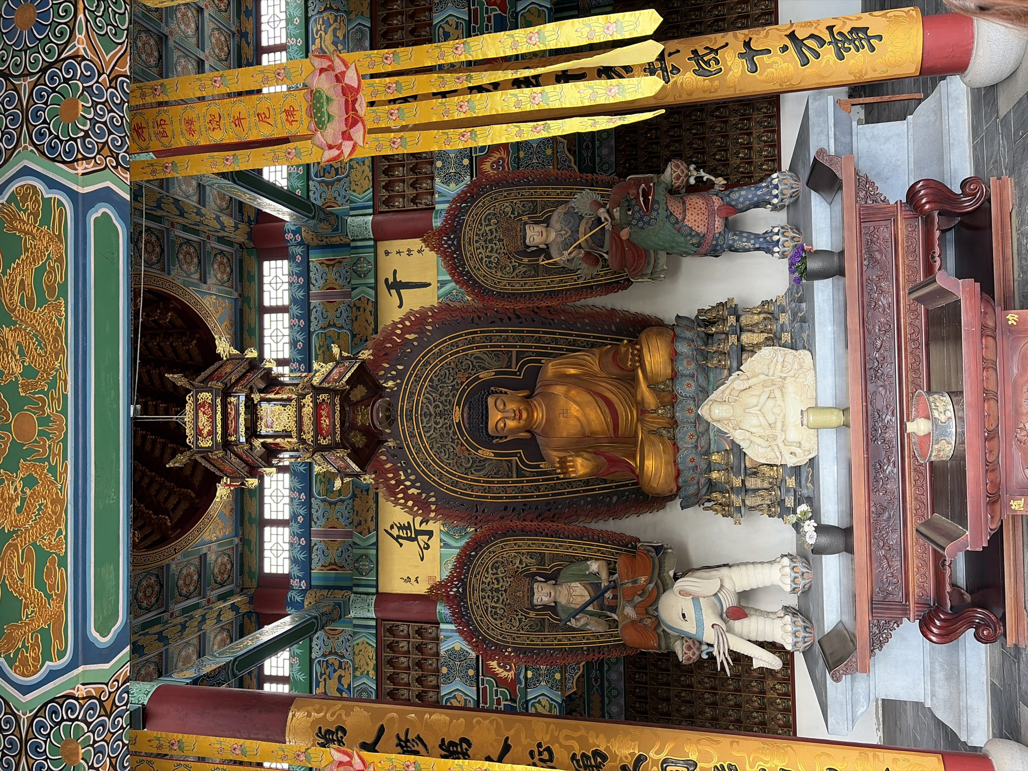 The image shows an altar within a Buddhist temple. A large, golden statue of Buddha sits at the center, flanked by two smaller statues on either side. The central Buddha statue is seated with its right hand raised in a blessing gesture. To the left of the central Buddha is a statue of a figure with a crown, seated on an elephant sculpture. To the right is another crowned figure, seated on a dragon-like sculpture. The altar is decorated with intricate carvings and ornate architectural elements. The ceiling above is adorned with colorful paintings of dragons and floral motifs. Yellow banners with calligraphic inscriptions hang from the ceiling. In the foreground, a wooden table with carved details is visible, holding a few decorative items. The overall impression is one of reverence and traditional Buddhist artistry. This scene is not located in Cambron-Casteau, Belgium.