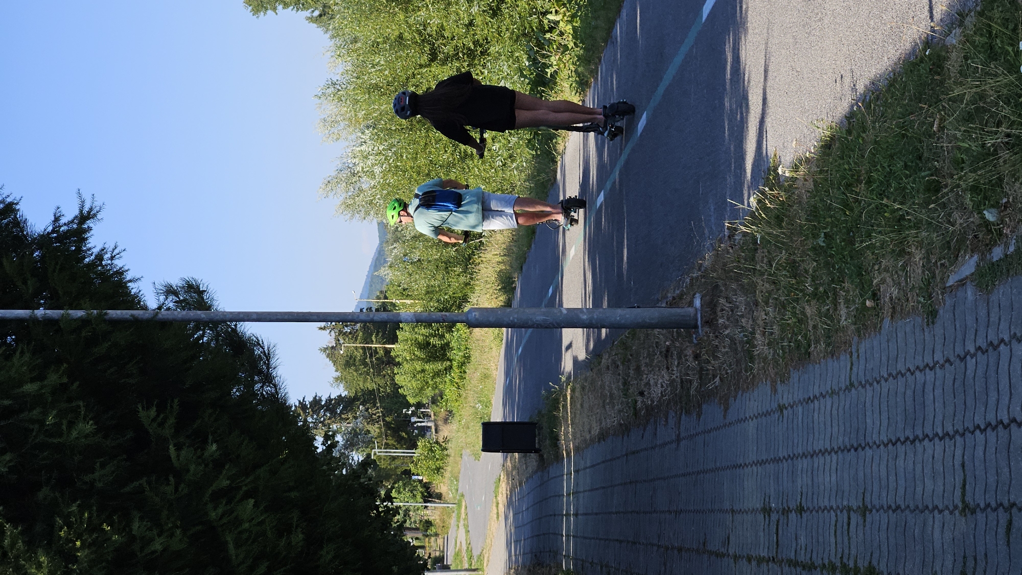 The image depicts a sunny day scene on a paved bike path in the city of Prešov, Slovakia. Two individuals are riding electric scooters on the path. The person in front, a male, wears a green helmet and a blue backpack, and is dressed in a light-colored t-shirt and shorts. The person behind, a female, wears a dark helmet and a black outfit, with visible legs. Both are moving away from the viewer. To the left of the bike path is a pedestrian walkway paved with grey interlocking bricks. A dark trash receptacle is visible near the edge of the pedestrian walkway. Lush green trees and vegetation line the right side of the bike path and the background, with a hill visible in the distance under a clear blue sky. A lamppost stands prominently on the right side of the path. The overall environment suggests a recreational or commuting setting within a park or along a thoroughfare.