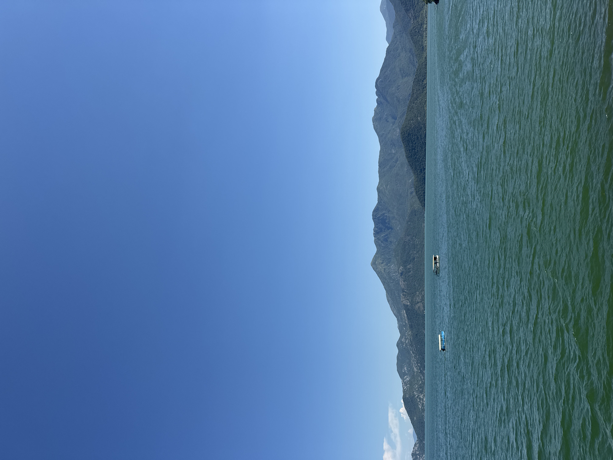 The image shows a view of Lake Piva, Montenegro. The clear blue sky occupies the left half of the image. On the right, a large body of water stretches from the bottom to the top, reflecting the sunlight and creating wavy patterns. In the center of the image, a mountain range rises along the edge of the lake. Two boats are visible on the water. The location is near Kruševica, Montenegro.