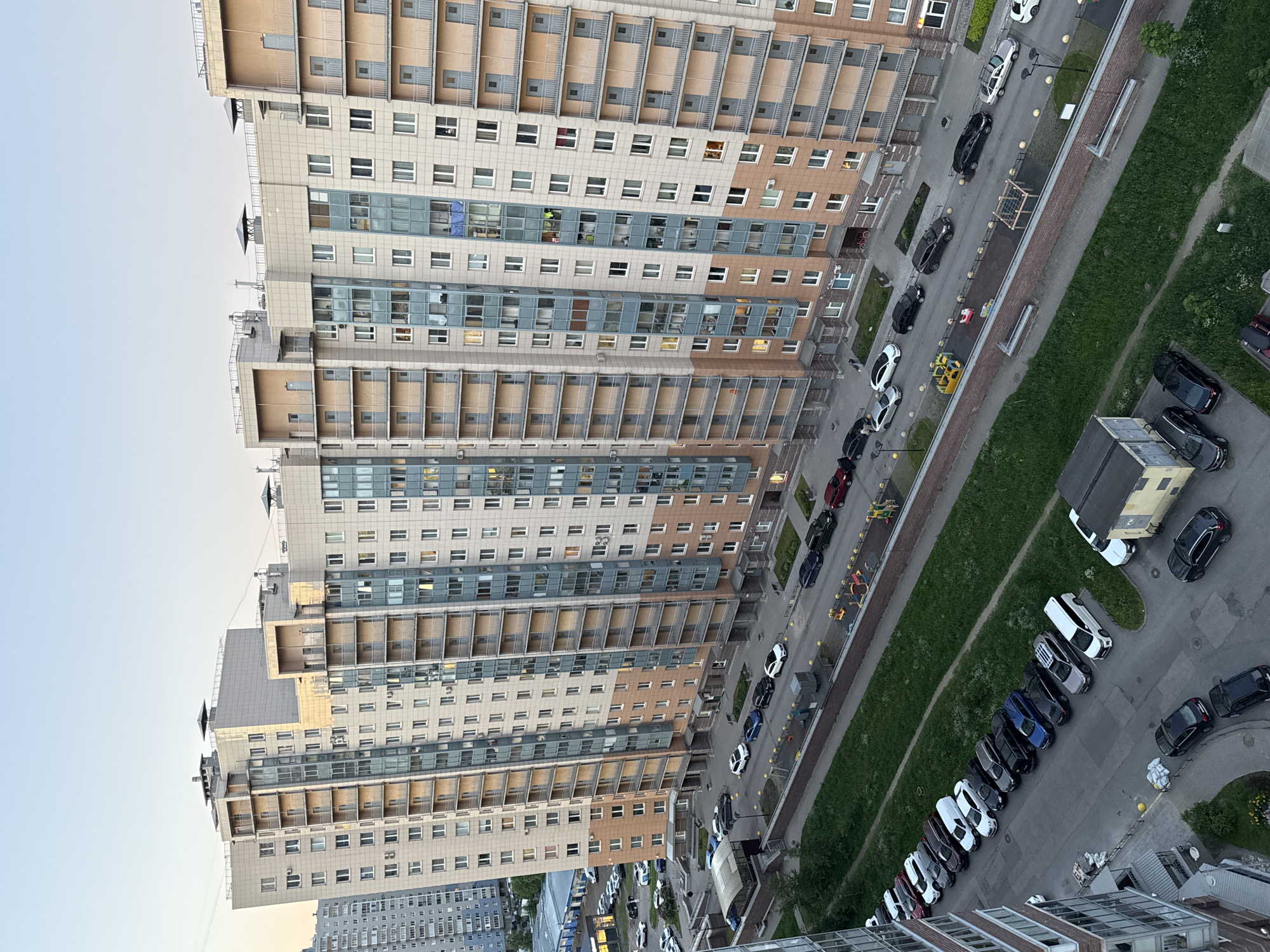 This high-angle urban landscape depicts a modern residential complex in an Unknown city, Russia, likely during an early morning or late afternoon with clear, pale blue skies. The dominant features are several tall, multi-story (approximately 15-20+ floors) residential apartment buildings forming a continuous block. Their facades are characterized by a modular design, primarily light beige or cream-colored panels, punctuated by vertical sections of darker blue-grey glass and metal, likely enclosing balconies or loggias. Some building sections, particularly on the right, feature peach-toned panels. Rectangular windows are numerous, with some showing internal illumination, suggesting occupancy. Visible on balconies are items such as a blue tarp in one window and what appears to be laundry in another. The flat rooftops are equipped with various utility structures, vents, and several decorative or functional pyramid-shaped covers. The rightmost building displays the Cyrillic characters "П" and "3" on its facade, indicating building numbers or sections. In the mid-ground to the left, a different, older-style multi-story grey residential building is visible, contrasting with the main complex. Below this, a light blue, low-rise commercial or service building with a flat roof and a partially visible red sign is present, surrounded by parked cars. The ground level features an intricate network of asphalt roads and parking areas. In front of the main buildings, vehicles are parked diagonally along curbs, and additional cars are in perpendicular spots. A long, narrow grassy strip separates this main forecourt from a lower road where a substantial number of cars, including sedans, SUVs, and vans (many dark-colored), are parked parallel to the curb. Further down in the lower-left, more vehicles are parked in an informal lot. A prominent white van, resembling a Mercedes-Benz Sprinter, is visible in the lower center-right. Pedestrian infrastructure includes sidewalks along the buildings and roads, marked by yellow bollards in places. Streetlights are positioned along the paths. A low brick wall topped with a metal fence separates some of the green areas from paved surfaces. Several small children's playgrounds are situated on a paved strip between the main buildings and the lower grassy area, featuring colorful equipment such as yellow swings, slides, and a wooden climbing structure. Sparse human activity is observed: one individual walks on a sidewalk in the mid-left, another is near a parked car in the lower-left, and a few indistinct figures are in the playground area. A red bicycle is parked near an entrance of