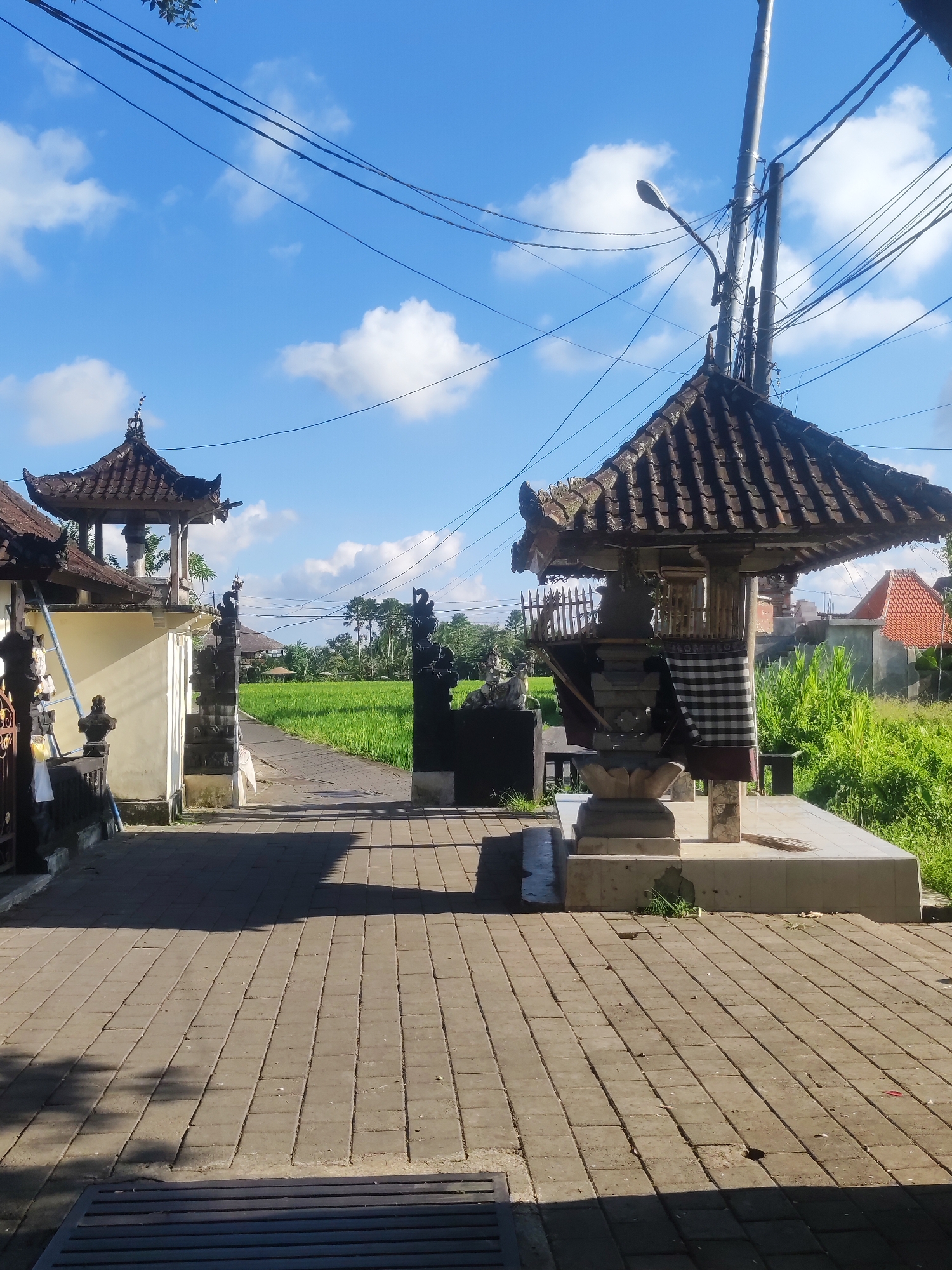 The image shows an outdoor scene in Sayan, Indonesia. A paved walkway leads through an entrance marked by traditional Balinese structures. On the right, a shrine with a tiled roof is adorned with a black and white checkered cloth. A similar structure is visible on the left, marking the entry. Beyond the entrance, a path stretches through a vibrant green rice field. Power lines cross the clear blue sky with scattered white clouds. A utility pole with a street lamp stands near the shrine on the right. Lush vegetation and glimpses of buildings can be seen in the background. The scene appears to be a quiet, rural setting within the city.