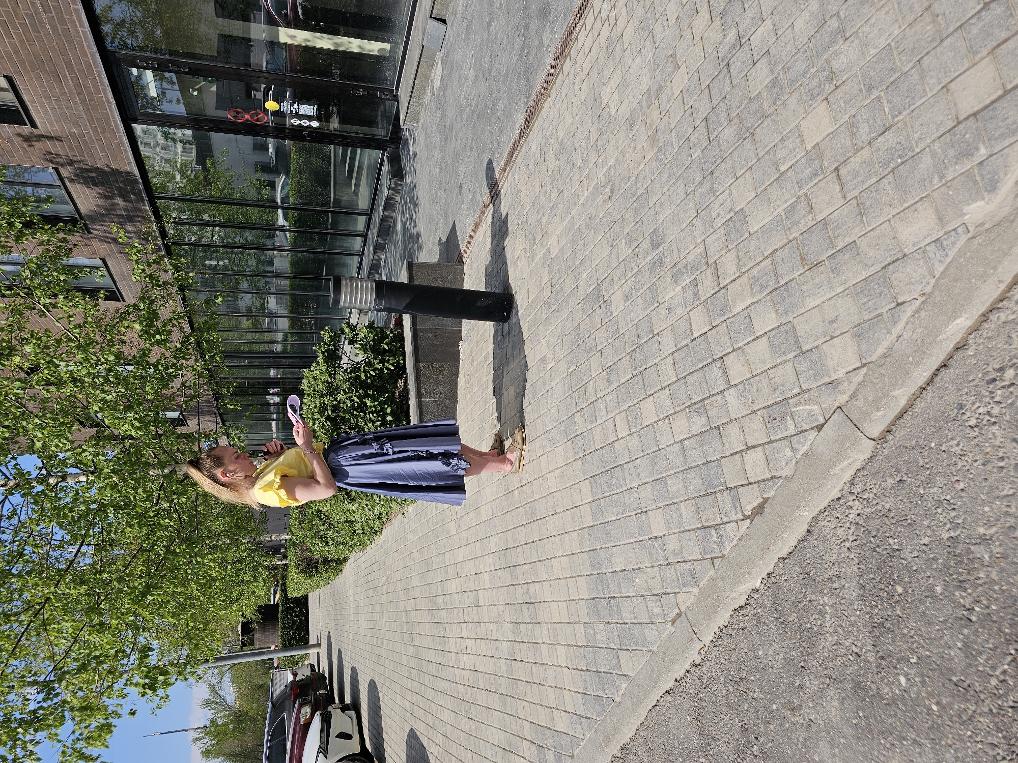 A woman with blonde hair stands on a brick-paved sidewalk in what appears to be a city environment. She wears a yellow blouse and a long, dark blue skirt, and is holding a small, white object (possibly a mirror or phone). She is looking at the object. The buildings along the left side have glass windows and brick facades. A tree is visible next to the woman. Parked cars are nearby and a tall, metal structure is visible in the background. Based on the architecture and style, the location could be in Moscow, Russia.