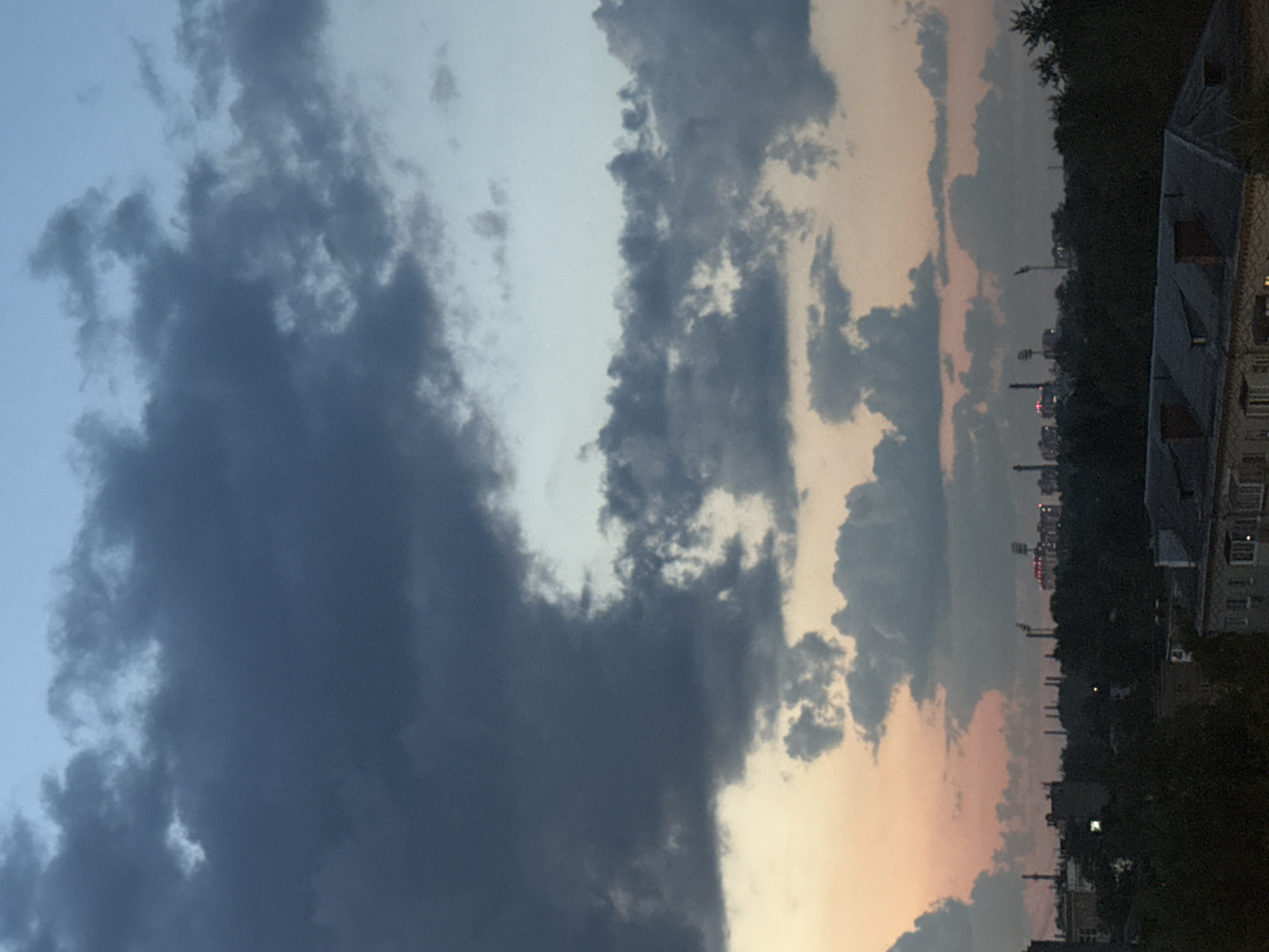 The image shows a city skyline at dusk, with a predominantly gray and blue sky filled with scattered clouds. The lower portion of the image features silhouettes of buildings and trees against the darkening sky. Some structures in the distance appear to be industrial, with lights visible on some of them. In the foreground, the rooftops of several buildings are visible, suggesting an urban residential area. The overall impression is of a slightly overcast evening in a city, possibly with industrial elements present. The scene captures the transition from day to night.