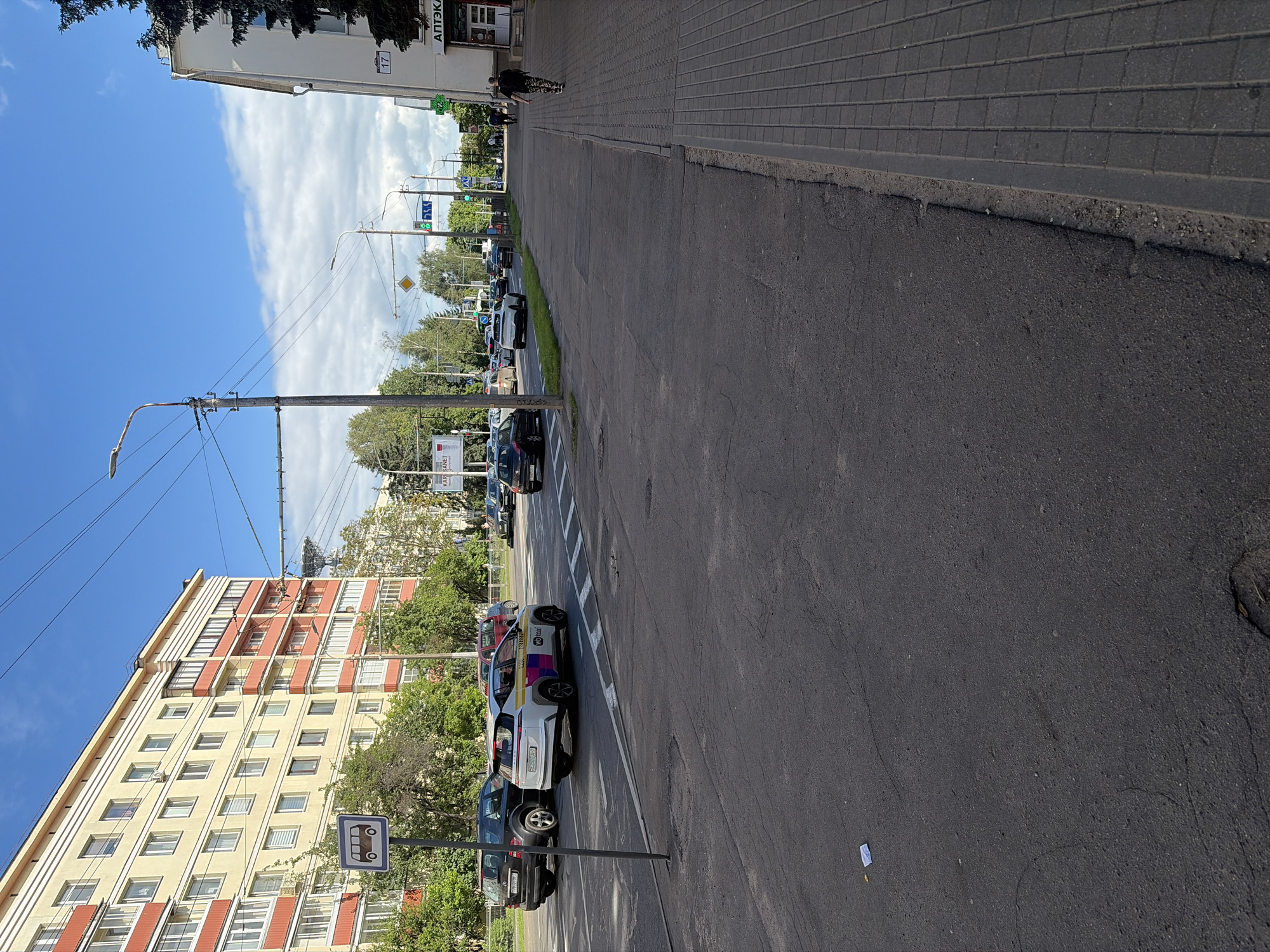 The image depicts a street scene in Minsk, Belarus, on a sunny day with a partly cloudy sky. The perspective is from the sidewalk, looking down a street lined with apartment buildings and trees.
In the foreground, a paved sidewalk runs alongside an asphalt road. A lamppost is visible on the sidewalk, with wires stretching across the frame. Several cars are parked or driving on the road. One car, a white taxi with colorful stripes and "VozTaxi" text, stands out. Other vehicles include dark sedans and SUVs.
On the left side of the street, a multi-story apartment building with light-colored walls and red balconies is prominent. Trees and greenery are interspersed along the street and in front of the building.
In the background, more apartment buildings, trees, and traffic lights are visible. There are also signs, including what appears to be a bus stop sign with a bus icon.
One building in the background has the text "АТТЭК" visible, likely indicating a pharmacy or similar establishment. The overall impression is of an urban environment during daylight hours, with typical street life.
