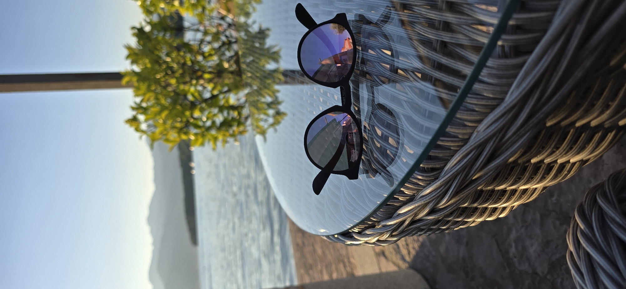 The image presents a pair of black, round-framed sunglasses lying on a clear glass table, reflecting the scene. The table has a woven base, and the backdrop features a body of water, likely the Adriatic Sea, with mountainous terrain in the distance. A small tree or bush with bright green foliage is partially visible in the upper left. The light suggests it is daytime. The reflection in the sunglasses suggests a building is present on the right side of the image, which is located in the city of Bogišići, Montenegro.
