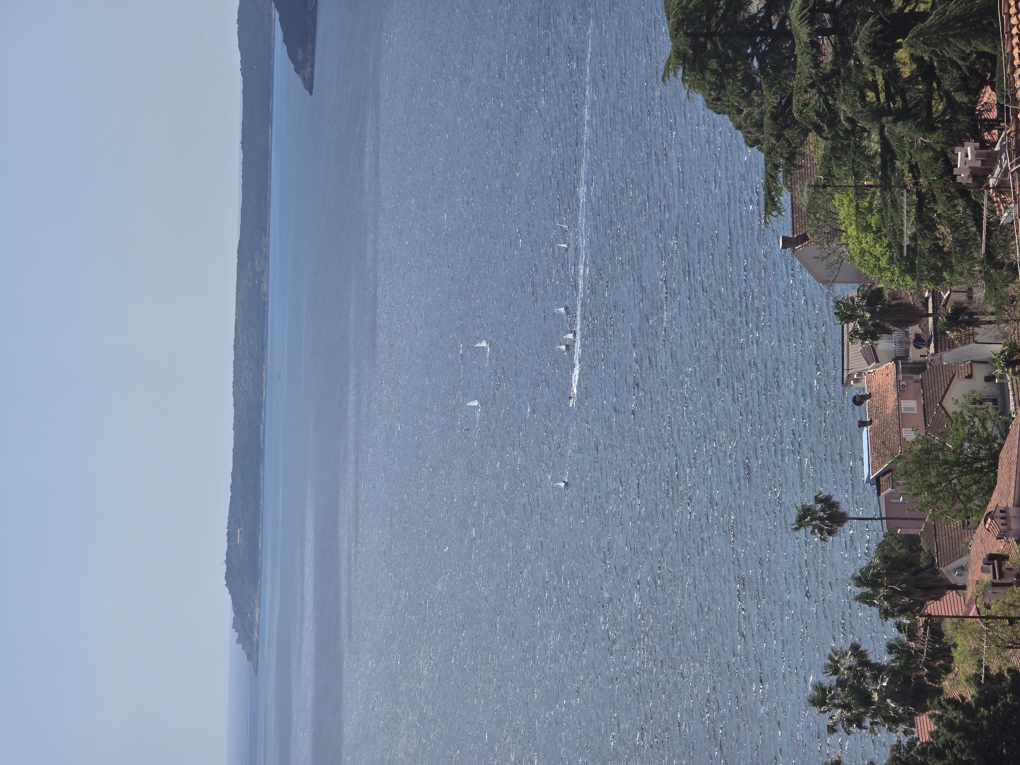 The image shows a panoramic view of the Adriatic Sea from Herceg Novi, Montenegro. Sailboats are scattered on the water. Buildings with red-tiled roofs are visible on the coastline. Trees and lush vegetation are interspersed with the buildings. A long, thin peninsula extends into the sea on the opposite side. The sky is light blue with a slight haze.