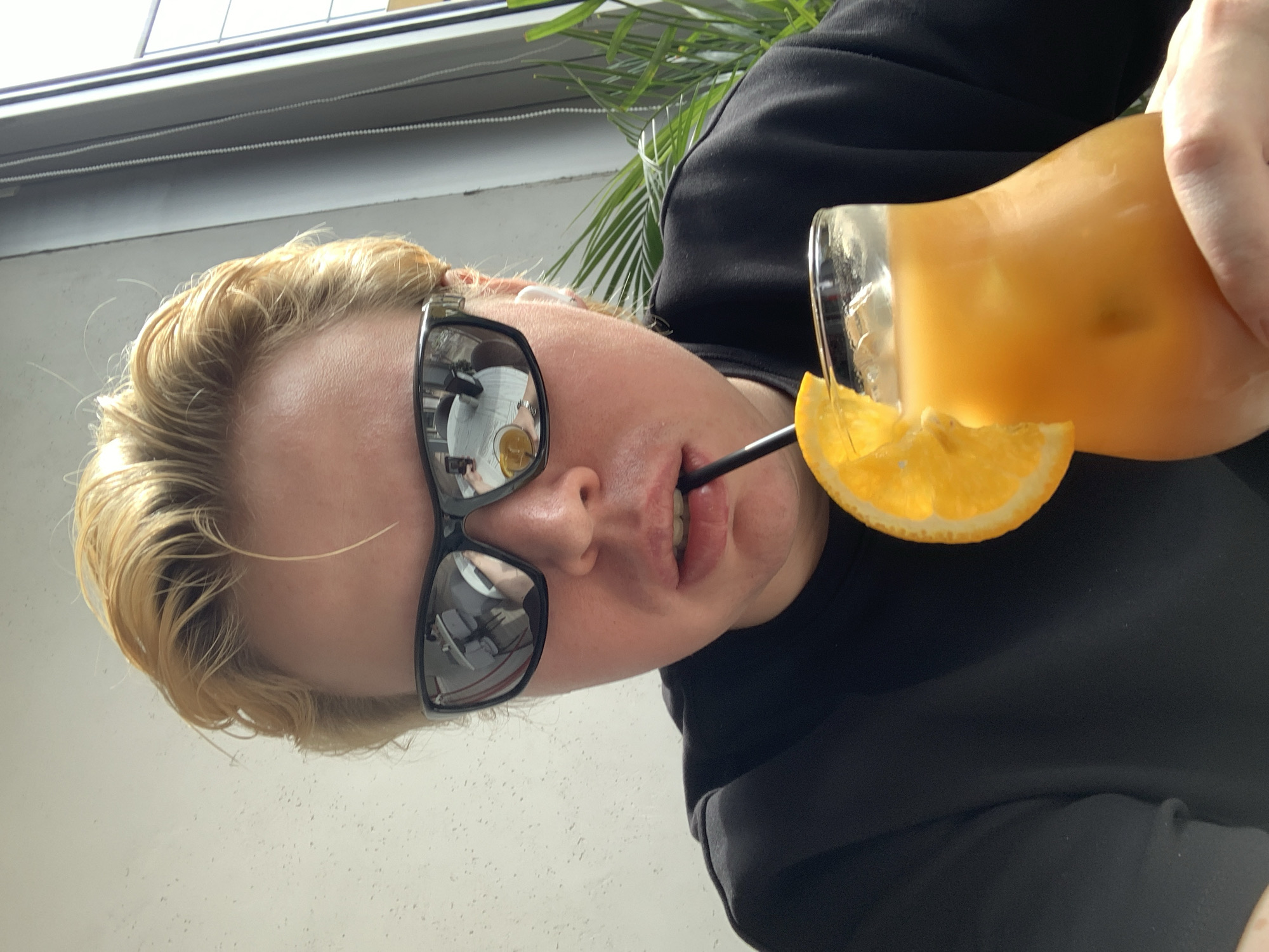 A person is holding an orange drink with a straw in their mouth. They are wearing sunglasses that reflect a table setting and a white shirt. The person is wearing a black top. The background includes a window, a white wall, and a green leafy plant. Based on the reflection in the sunglasses, the location might be a cafe or restaurant in Almaty, Kazakhstan.