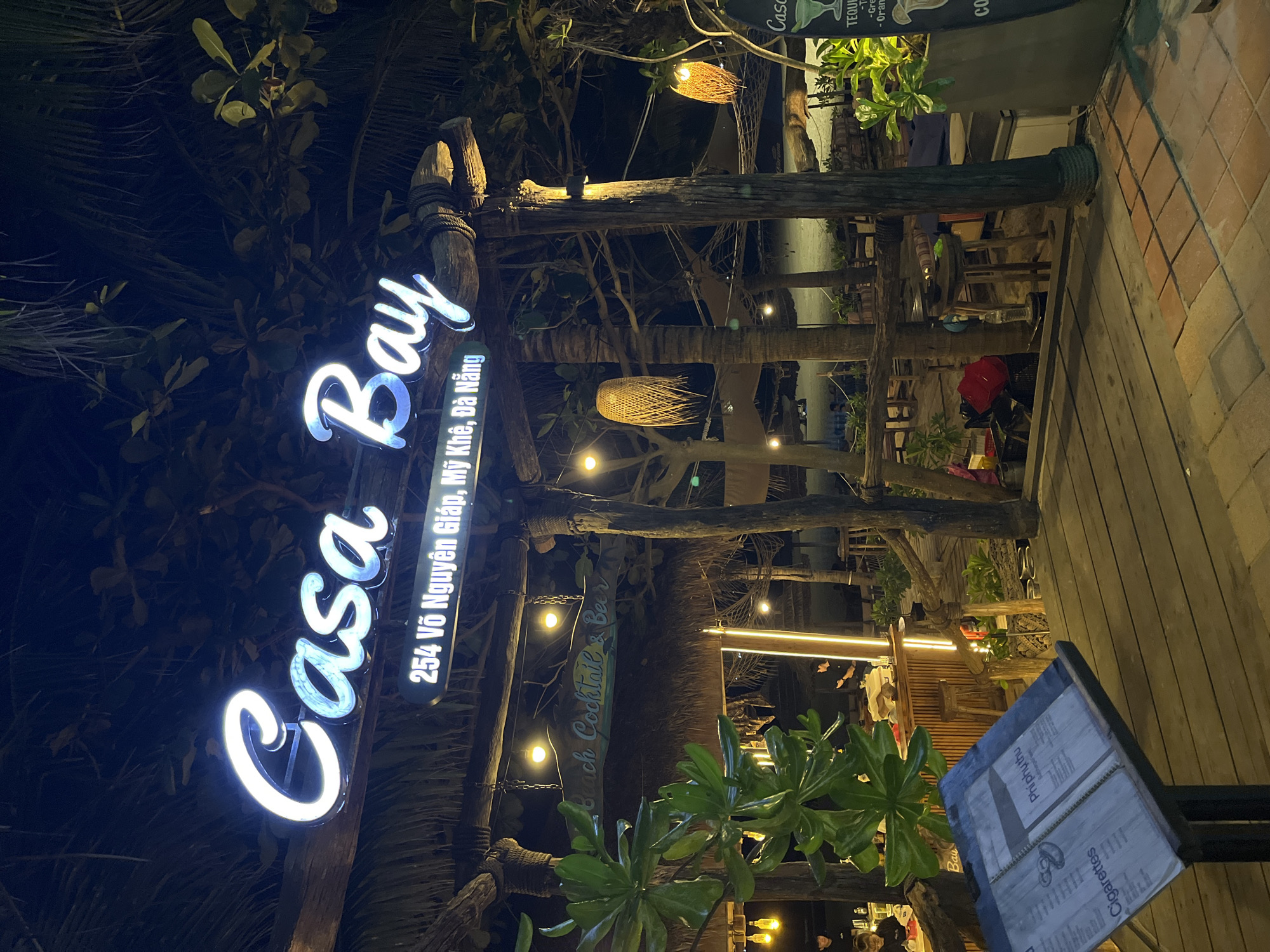 Here's a detailed description of the image:
The image presents an evening scene at "Casa Bay", a beachside establishment in Đà Nẵng, Vietnam, specifically located at 254 Võ Nguyên Giáp, Mỹ Khê. The name "Casa Bay" is prominently displayed in glowing, neon-like cursive lettering on a rustic wooden structure. Adjacent to the main sign, there is a smaller sign indicating "254 Võ Nguyên Giáp, Mỹ Khê, Đà Nẵng".
The scene is illuminated by string lights hanging from the wooden beams and thatched roofing of the structure. There are also decorative woven lanterns interspersed among the lights. In the foreground, a menu stand is visible, featuring options like "Cigarettes" and "Phí phụ thu".
The establishment is decorated with tropical foliage. The scene suggests a beach bar setting, with the backdrop showing a glimpse of the ocean and beach. The overall atmosphere appears relaxed and inviting. Patrons are vaguely visible in the background, suggesting active business activity.
Additionally, another sign reads "Beach Cocktail & Beer," reinforcing the identity of the venue. A vertical sign containing "Casa" is visible, along with drinks information, including "Tequila".
