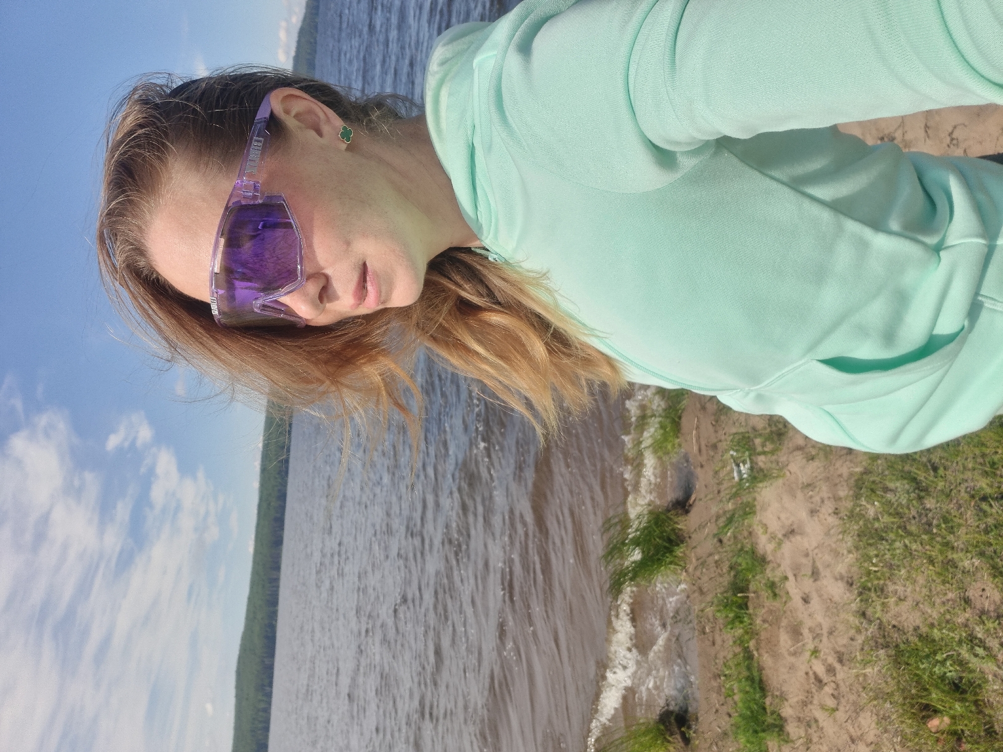 A person is depicted in an angled, close-up shot, positioned outdoors beside a large body of water. The subject, appearing to be female, has long, light brown hair and is looking slightly downwards. She wears striking purple, wrap-around sunglasses that obscure her eyes, and a small, green clover-shaped earring in her right ear. Her attire consists of a light mint green or aqua-colored hooded sweatshirt.
The background features an expansive body of water, likely a lake or a wide river, with small waves gently reaching the shore. Across the water in the distance, a dark green treeline defines the opposing bank. The sky above is clear and blue with scattered white clouds, indicating sunny daytime weather. The foreground shows a sandy or dirt bank interspersed with patches of green grass.
The scene suggests a serene, contemplative moment spent outdoors, possibly by the water's edge. Faint text, which appears to be "BLIZ," is visible on the arm of the purple sunglasses. Given the provided context, the location is Chaikovsky, Russia, fitting the description of a natural waterside setting.