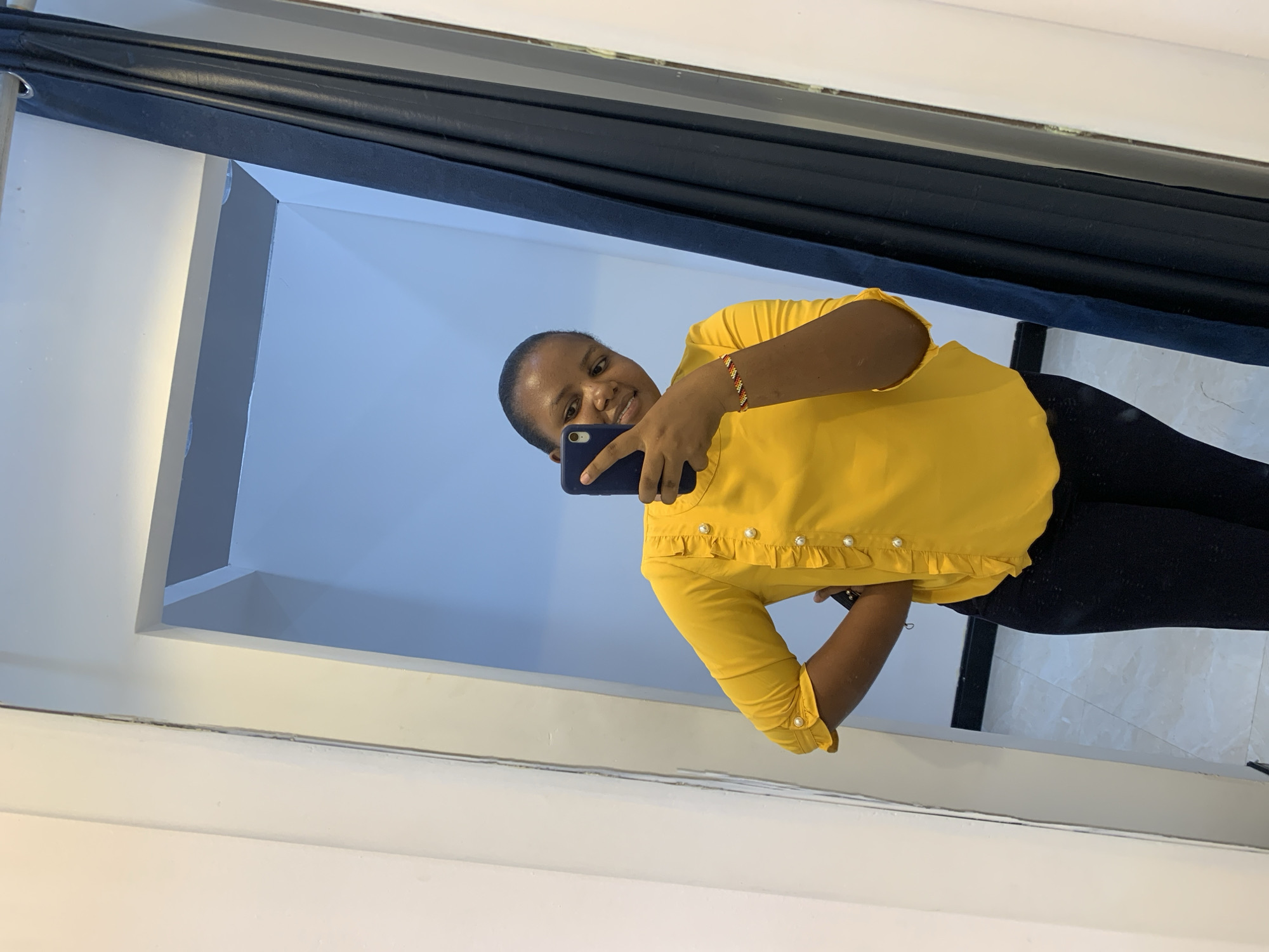 A woman is reflected in a mirror, holding a smartphone and taking a selfie. She is wearing a yellow blouse with ruffles and pearl-like buttons, and black leggings. The background suggests an indoor space with white walls and a dark curtain or drape visible near the top of the frame. The exact location in Dar es-Salaam, Tanzania is not discernible from the image.
