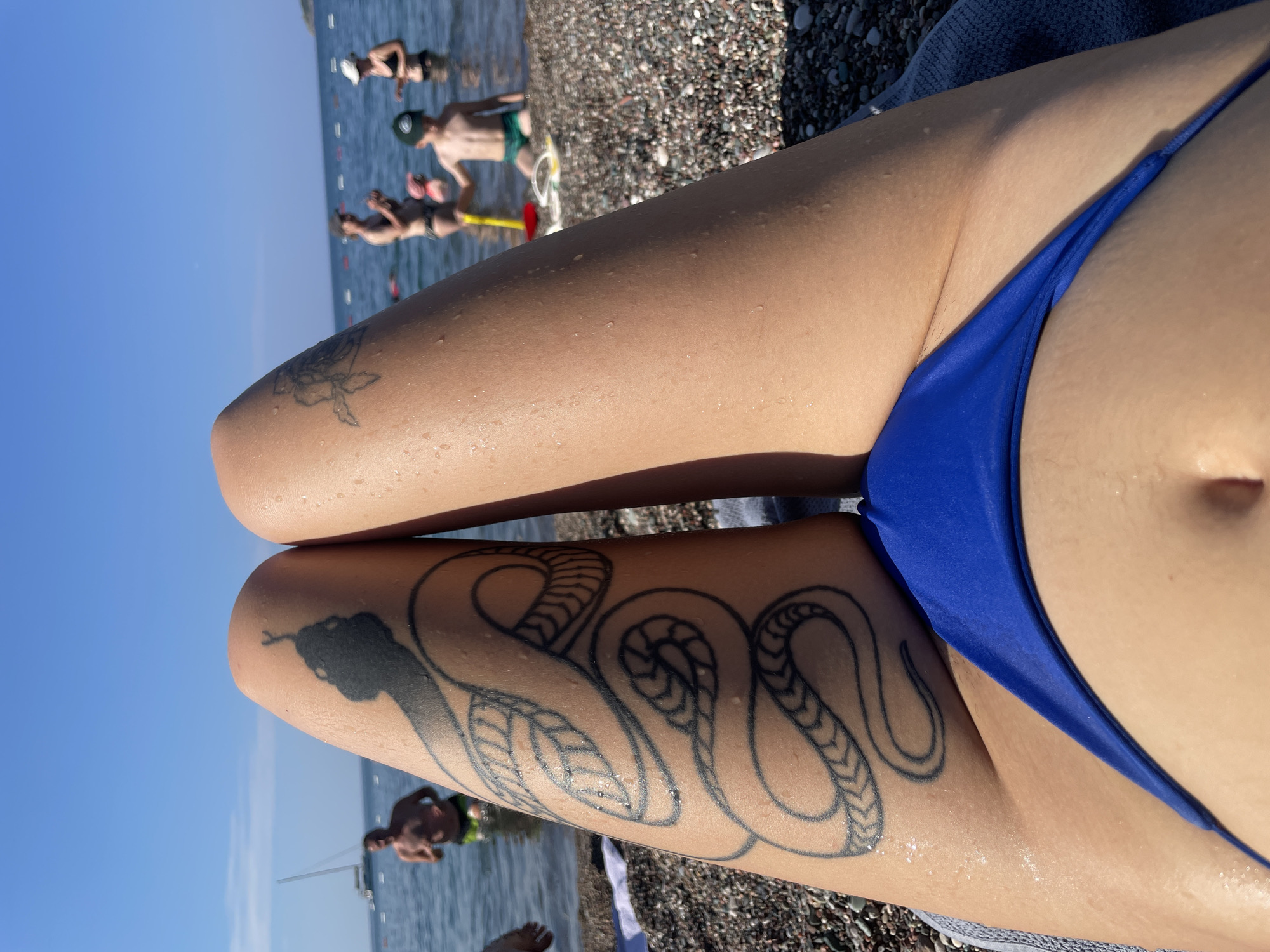 The image shows a close-up view of a person's legs at a beach in Rafailovići, Montenegro. The person has a snake tattoo on their left thigh and is wearing a blue bikini bottom. In the background, several people are in the shallow water, including adults and a child playing with a shovel and bucket. A sailboat is visible on the water in the distance. The beach is covered in pebbles. The sky is clear and blue, indicating a sunny day.