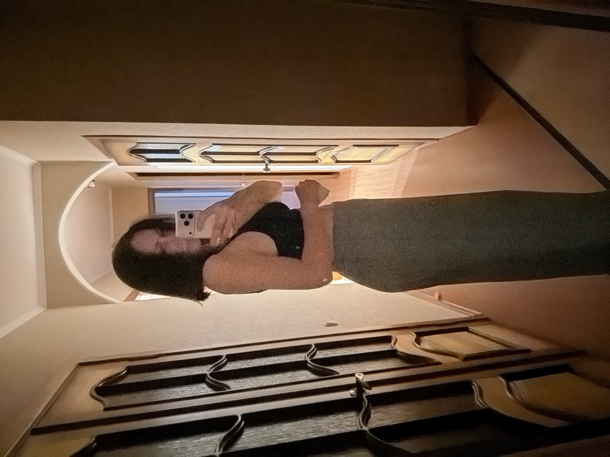 A woman stands in front of a mirror taking a selfie. She is wearing a black top and gray leggings. The mirror is framed by a dark brown wooden frame. The walls are light beige. The ceiling is dark brown. The location is Unknown, Unknown.