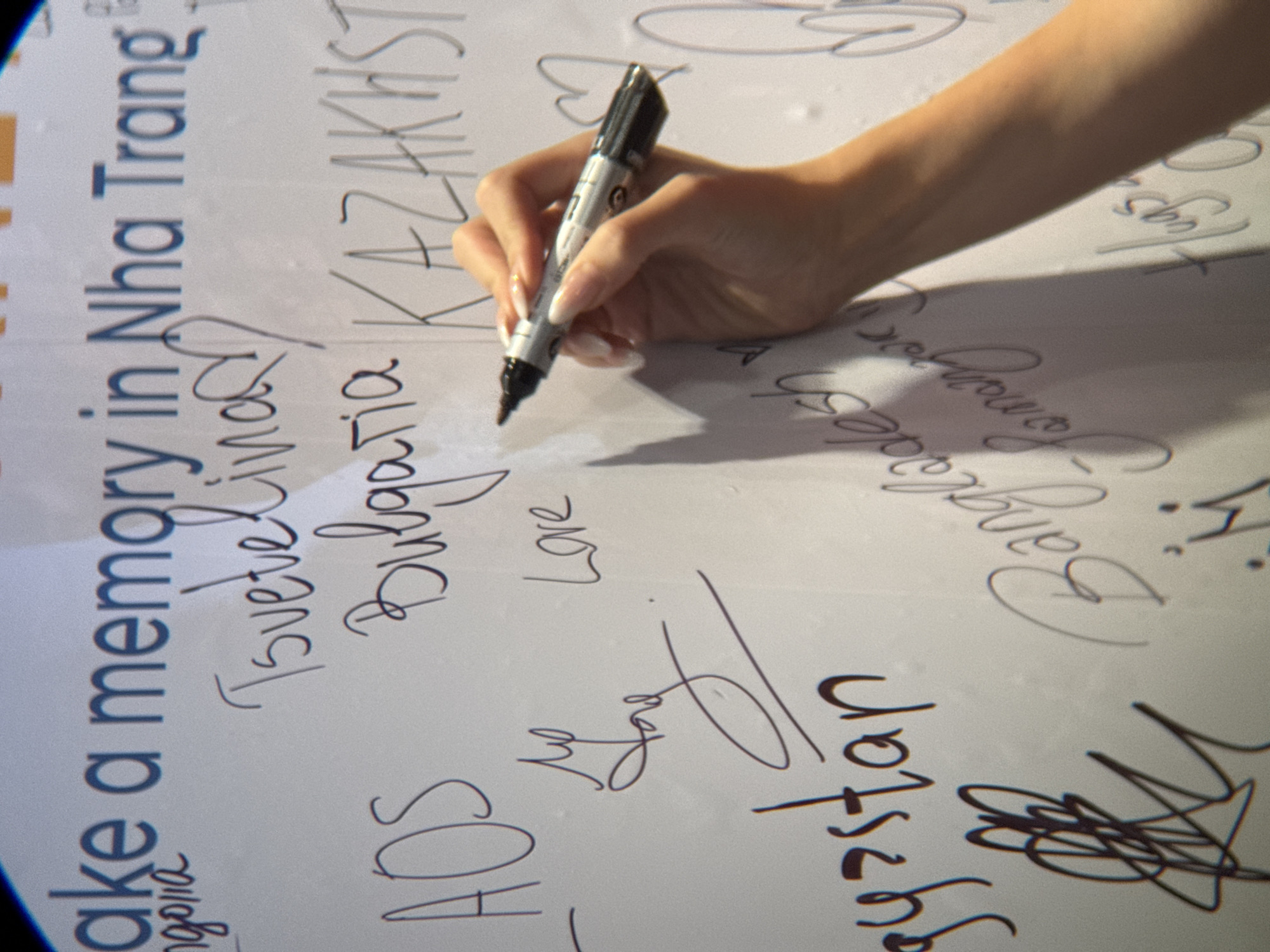 A close-up shot captures a person's hand holding a black marker and writing on a whiteboard. The marker is positioned to the right of the frame, with the hand holding it and the tip of the marker poised to write on the surface. The whiteboard displays various handwritten messages, names, and possibly locations. Above the writing, a blue text reads "ake a memory in Nha Trang," suggesting the location is Nha Trang, Vietnam. Other visible handwritten text includes names like "Tsvetelinav," "Bulgaria," "KAZAKHSTAN," "AOS," "gyzstan," "Bangladesh," and "Hugs." The overall environment appears to be indoors, with the whiteboard being the primary subject in the foreground. The lighting casts a shadow from the hand and marker onto the whiteboard.