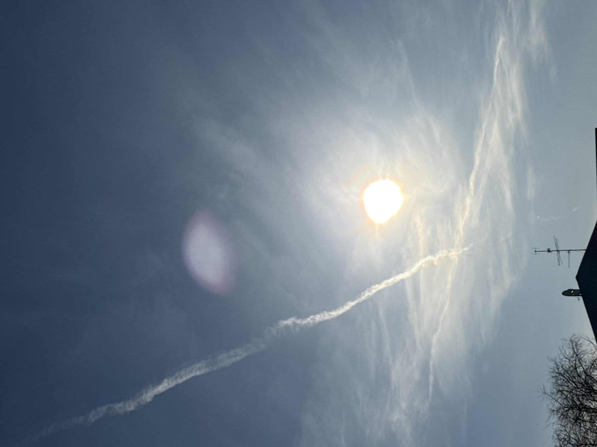 The image shows a daytime sky with the sun visible through thin, wispy clouds. A prominent contrail stretches diagonally across the frame, appearing to originate from behind the sun. A lens flare is visible to the left of the sun. In the lower right corner, a partial view of a building is seen, including a television antenna and bare tree branches. The location of the image is unknown, since there is no enough information to provide specific city information.