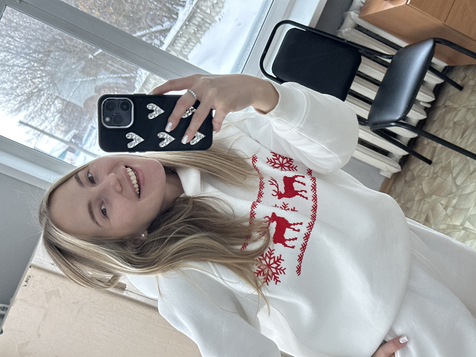 A female subject is captured in a selfie taken indoors, appearing to be reflected in a mirror or glass surface. She has long, blonde hair and is smiling broadly, revealing her teeth. She wears a white hooded sweatshirt adorned with a red Nordic-style pattern featuring two deer and snowflakes across the chest, along with matching white sweatpants. A small stud earring is visible in her left ear. Her left hand holds a black smartphone with a case decorated with seven silver heart-shaped embellishments; a ring with small stones is visible on her left ring finger, and her nails have white tips.
The indoor setting includes a large window on the right, providing a view of a snow-covered outdoor environment with bare trees and indistinct distant structures. Below the window, a white fluted radiator is visible. A black metal-framed chair with a black padded seat and backrest is positioned in the lower right foreground. The floor appears to be a light-colored wood or laminate. A segment of a light brown wooden furniture piece is visible at the bottom right corner. The left side of the indoor space shows a plain, light-colored wall. The scene indicates a winter day in an unknown indoor location within an unknown city.