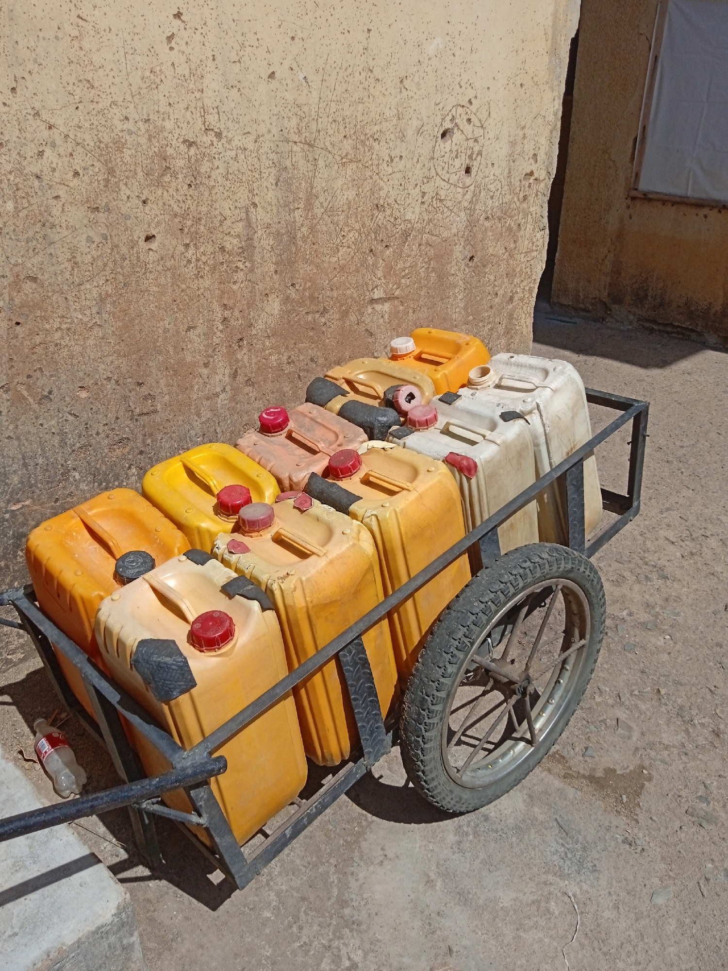 A metal cart filled with nine plastic containers sits outdoors on a concrete surface. The containers are mostly yellow, with some orange and white ones visible. The cart has a single tire with a worn tread. A discarded plastic bottle rests near the cart. The setting appears to be a street or alleyway, with a beige stucco wall forming the backdrop. Based on the visual clues, the location is likely in Jos, Nigeria.