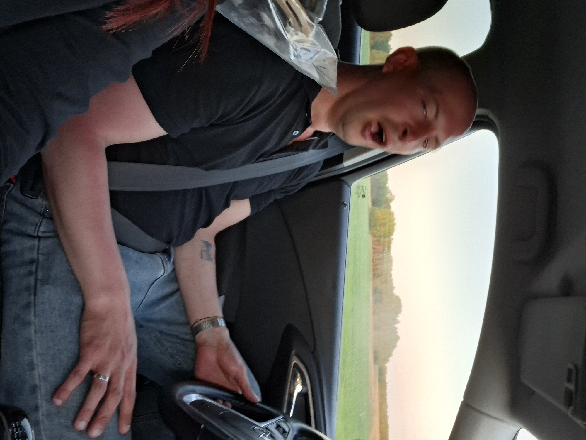 A man is seated in the driver's seat of a car, wearing a black shirt and light blue jeans. He is also wearing a seatbelt. His mouth is open, possibly talking or yawning. He has a wedding ring on his left ring finger and a bracelet on his left wrist. A tattoo is visible on his left forearm. The car's interior is visible, including the steering wheel and dashboard. Outside the window, there is a field, some trees, and a light-colored sky. The location appears to be the city of Любанскі сельскі Савет, Belarus, judging by the landscape and sky visible through the car window.