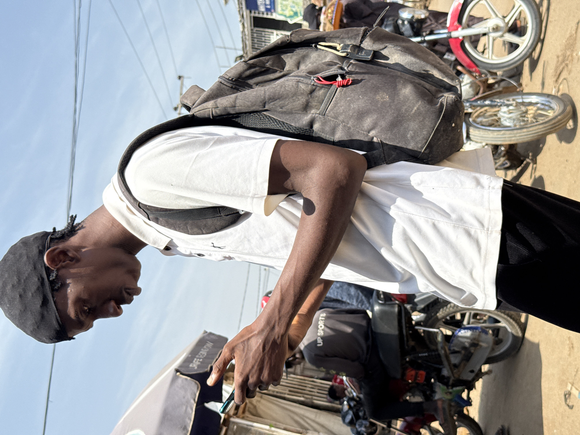 A young Black male, wearing a black skullcap, a white polo shirt, and black pants, is standing in an outdoor marketplace in Abuja, Nigeria, looking down at his smartphone. He has a large, dark gray backpack slung over his right shoulder, its straps taut. His hands are holding the phone, which is emitting a faint green glow. The background is a blur of activity, with several motorcycles parked nearby. One motorcycle in the foreground has a red and silver body. Behind the man, another person is visible wearing a gray shirt with "UP SPORTS" printed on the back. The sky above is clear and bright blue, with a few thin power lines crisscrossing it. The ground appears to be dirt or gravel. A portion of a grey and white awning is visible to the left of the man.