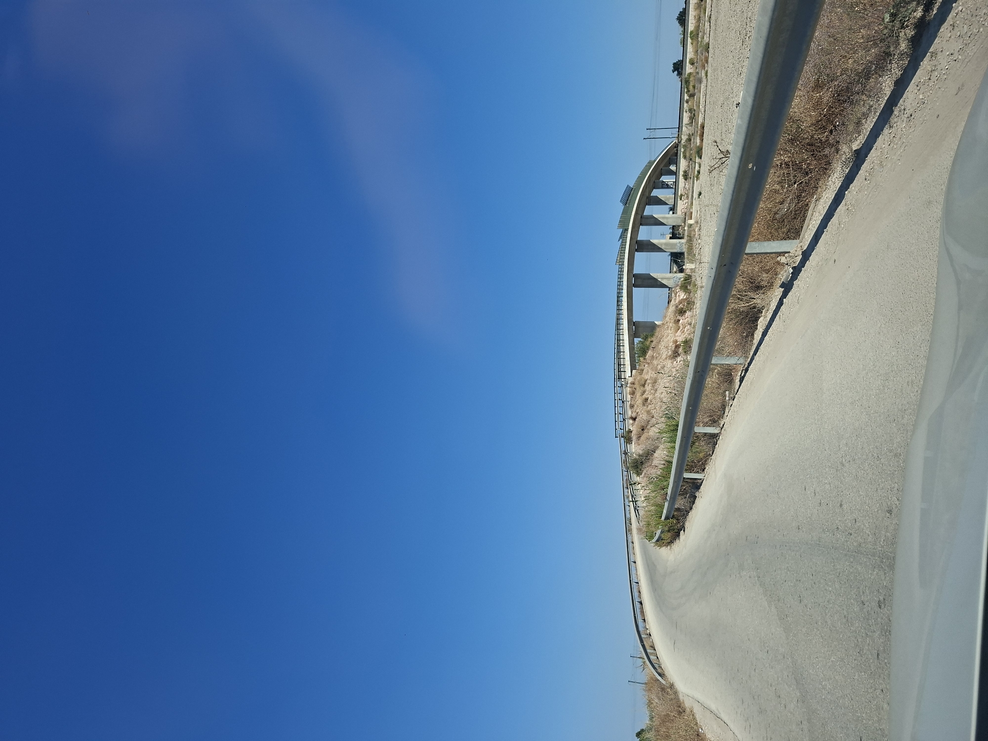 A wide, curved, paved road ascends towards a modern concrete overpass under a clear, bright blue sky. The road is bordered by a silver metal guardrail on the right and dry, sparse vegetation on the left. The overpass, with its arched structure and multiple vertical supports, spans across the landscape. The terrain surrounding the road and under the overpass appears arid and dry, with patches of scrub and dirt. In the far distance, a few electrical poles are visible. The image seems to capture a scene from a vehicle, as indicated by the foreground which suggests a dashboard or hood. The location is identified as Callosa de Segura, Spain.