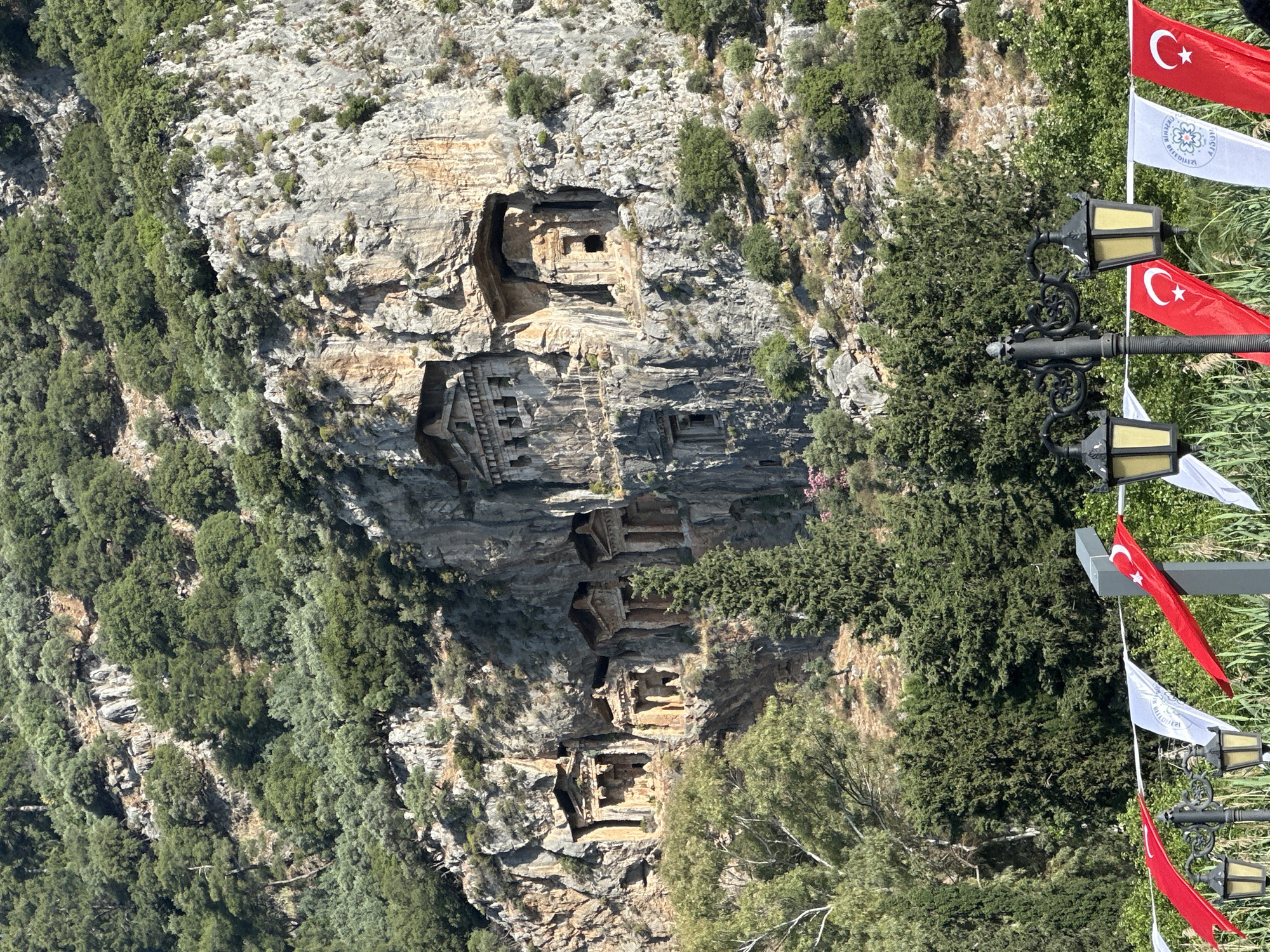 The image captures the ancient Lycian rock tombs in Dalyan, Ortaca, Turkey, carved into a cliff face overlooking the Dalyan River. The tombs are multi-tiered and have ornate facades. Lush greenery covers the cliff edges around the tombs. In the foreground, there are lamp posts decorated with Turkish flags and white banners.