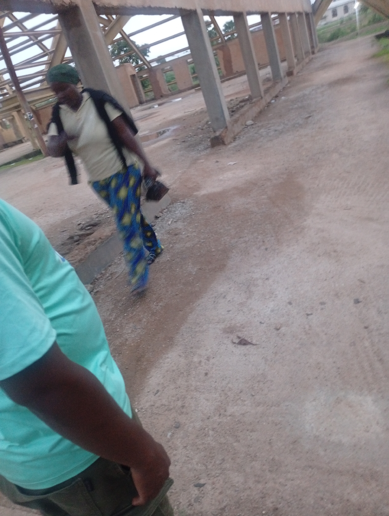 The image shows an outdoor scene in what appears to be a construction or unfinished building site. In the foreground, a person is partially visible, wearing a light blue t-shirt and olive green pants. Their arm is extended towards the viewer, with their hand in their pocket. In the midground, a woman is walking, wearing a green head covering, a light-colored t-shirt, a dark jacket draped over her shoulders, and brightly colored blue and yellow patterned pants. She is carrying a dark bag in her right hand. The environment features unpaved ground with scattered debris. The background shows the skeletal structure of a building with exposed beams and concrete pillars. The overall lighting suggests it may be overcast or late afternoon. The location is identified as Jos, Nigeria.