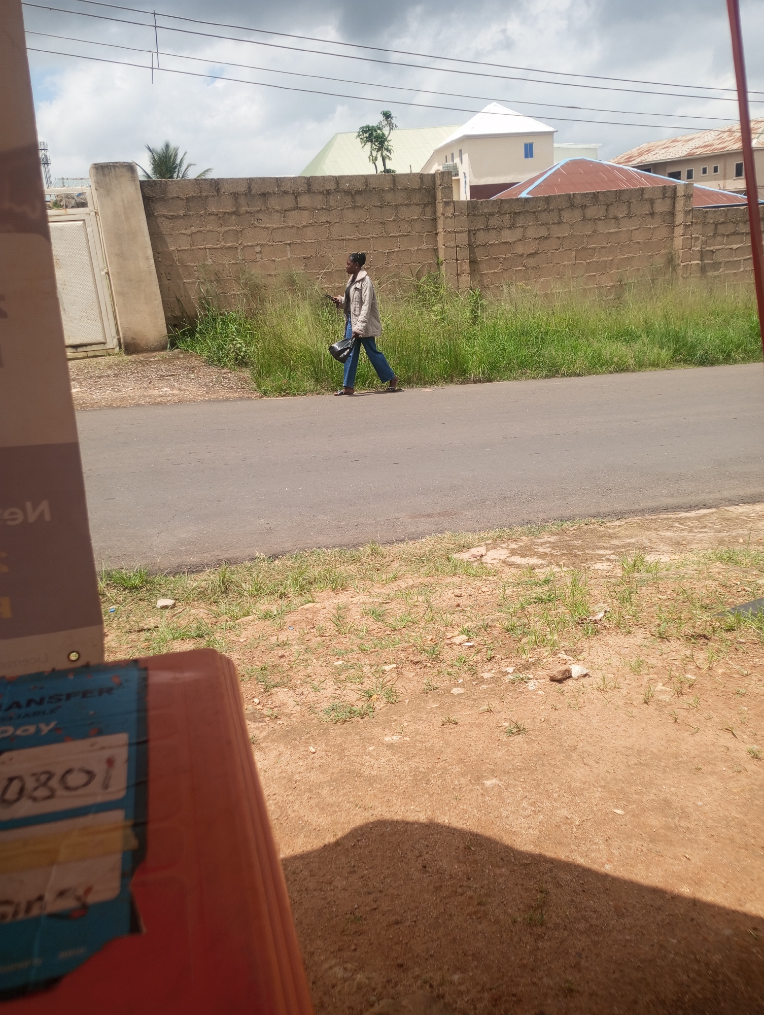A person walks along a paved road in Jos, Nigeria. The person is wearing a light-colored jacket and blue pants and is carrying a black bag. They are walking away from the camera and are on the right side of the road. Behind the person is a low, light brown brick wall with green grass growing in front of it. Beyond the wall, several buildings can be seen, including one with a white, pointed roof and another with a reddish-brown corrugated metal roof. The sky is partly cloudy. In the foreground, a portion of a red surface is visible, with some blue and white paper attached to it, featuring text that includes "TRANSFER" and numbers.