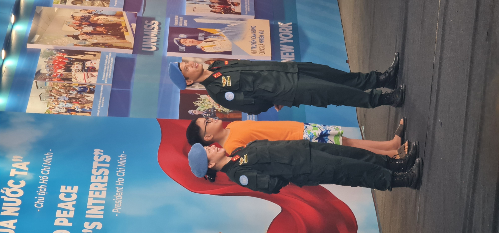 This media file captures an indoor scene, likely an exhibition or event in Đà Nẵng, Vietnam, centered around United Nations peacekeeping missions. Three individuals are prominently featured in front of a large informational display.
In the foreground, two individuals are dressed in dark green/black military uniforms, black combat boots, and blue berets, indicating their roles as UN peacekeepers. One is a male and the other a female, both standing formally. Between them is a civilian male, dressed in an orange T-shirt and white, blue, and green patterned shorts, wearing sandals. He is looking upwards towards the display.
The background consists of a large blue and white display wall adorned with text and photographs. Visible text in Vietnamese includes "JÀ NƯỚC TA" (Our Country), "Chủ tịch Hồ Chí Minh" (President Ho Chi Minh), and "KHI TRUYỀN CẢM HỨNG CŨNG LÀ NHIỆM VỤ" (When inspiring is also a duty). English text includes "PEACE", "'S INTERESTS'", "President Ho Chi Minh", "UNMISS" (United Nations Mission in South Sudan), "UNPOL" (United Nations Police), and "NEW YORK". Several photographs showing groups of people, possibly peacekeepers or mission activities, are also part of the display.
The overall scene suggests an educational or commemorative event highlighting Vietnam's contributions to UN peacekeeping efforts. The uniformed individuals appear serious and professional, while the civilian seems engaged with the exhibit. The lighting suggests an indoor setting, possibly an exhibition hall.